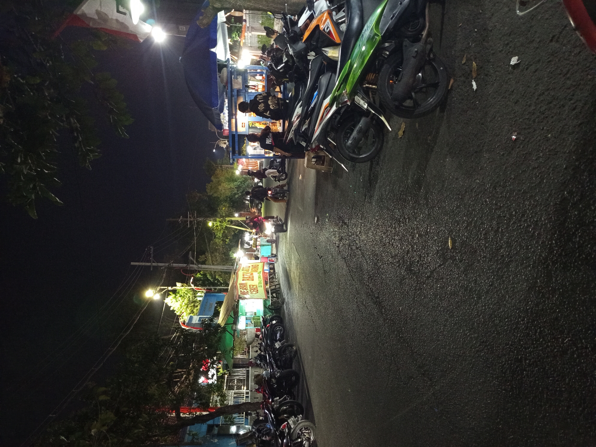 The image depicts a street scene at night in Sidoarjo, Indonesia, likely a commercial area with food vendors and a significant presence of parked motorcycles. The lighting is predominantly from streetlights and the illuminated stalls.
On the left side of the street, a row of parked motorcycles is visible, with some of them appearing to be in a designated parking area. Behind the motorcycles on the left, there is a blue building with a red and white flag or banner. Further down the street on the left, a food stall with a yellow banner advertising "AYAM BATU CEKER AYAM MOJING" is illuminated.
In the center of the image, several motorcycles are either parked or in motion. One motorcycle with a rider is moving towards the right. In the background, there are more food stalls with blue and white canopies, and people are visible interacting around them. One stall has a blue umbrella providing shade.
On the right side of the street, more motorcycles are parked, including a green scooter in the foreground. Two individuals are standing behind a stall with a blue canopy. The ground is asphalt and appears slightly wet, reflecting the lights. The overall environment suggests a lively evening street scene with food vendors catering to customers arriving on motorcycles.