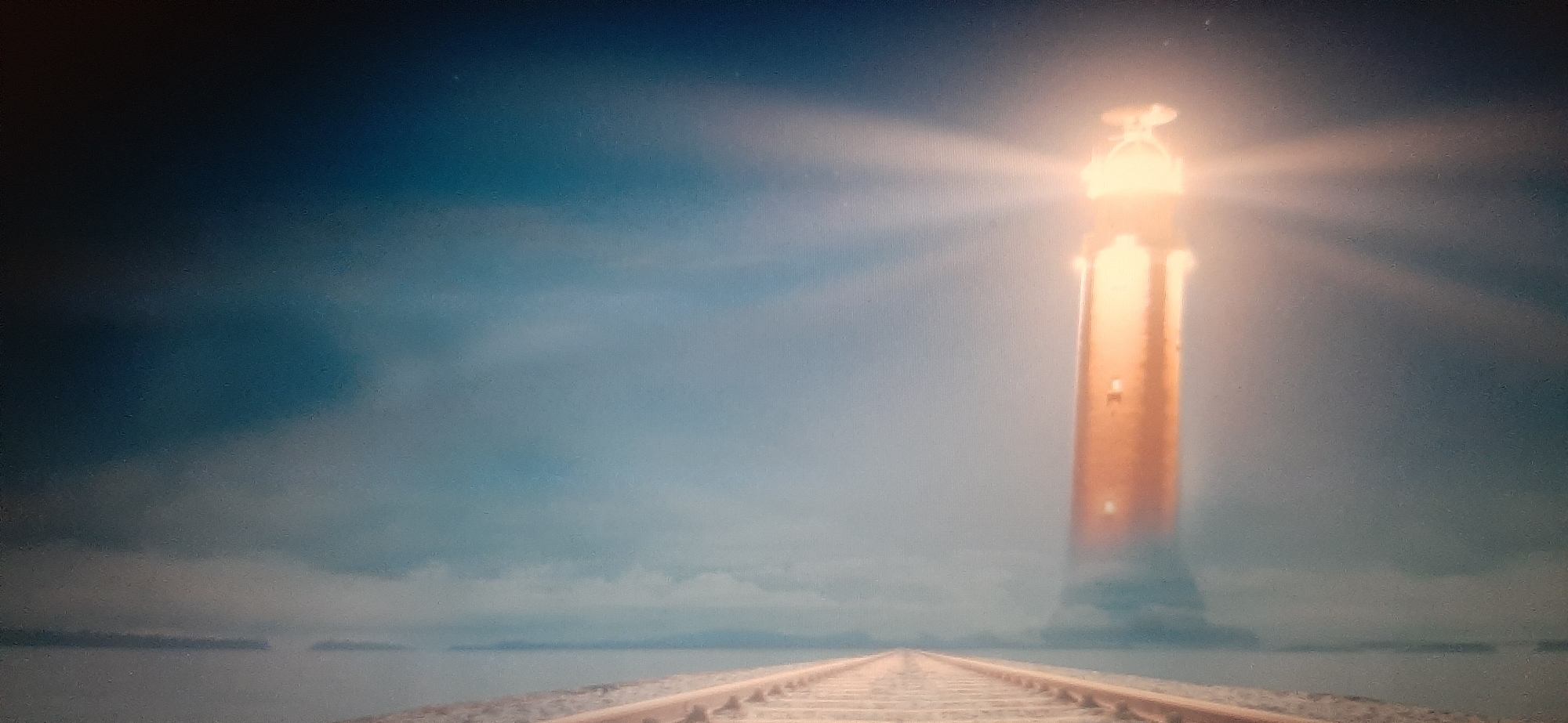 A surreal, digitally rendered image depicts a solitary lighthouse emitting a powerful beam of light across a misty, ethereal landscape. The lighthouse, rendered in warm golden tones, stands tall and imposing against a gradient sky that shifts from deep blue at the top to a lighter, hazy blue towards the horizon. The base of the lighthouse appears to merge with swirling mist or clouds, creating a sense of it emerging from another realm. In the foreground, a set of train tracks stretches out towards the lighthouse, disappearing into the atmospheric haze. The tracks are rendered in earthy tones, contrasting with the luminous quality of the lighthouse and sky. The overall scene evokes a feeling of mystery, guidance, and perhaps a journey towards an unknown destination. This image is not located in the city of Yessentukskaya, Russia.