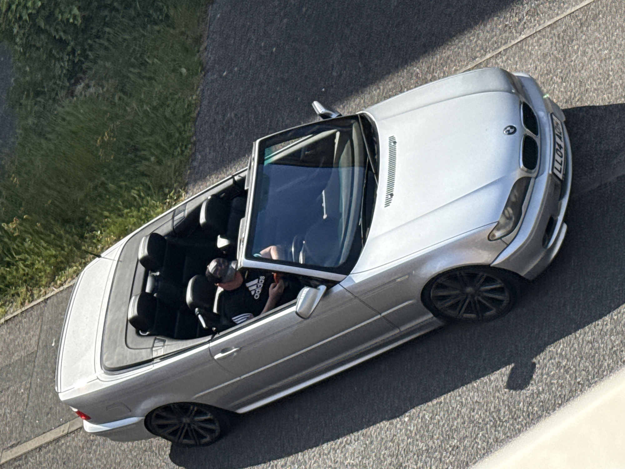 An aerial view shows a silver BMW convertible parked on a grey asphalt road. The car's top is down. Inside, a man wearing a baseball cap and a black Adidas t-shirt is visible in the driver's seat. The car is parked alongside a patch of green grass. The UK license plate LLO4 AOH is visible on the front of the car. This image was taken in Great Yarmouth, United Kingdom.