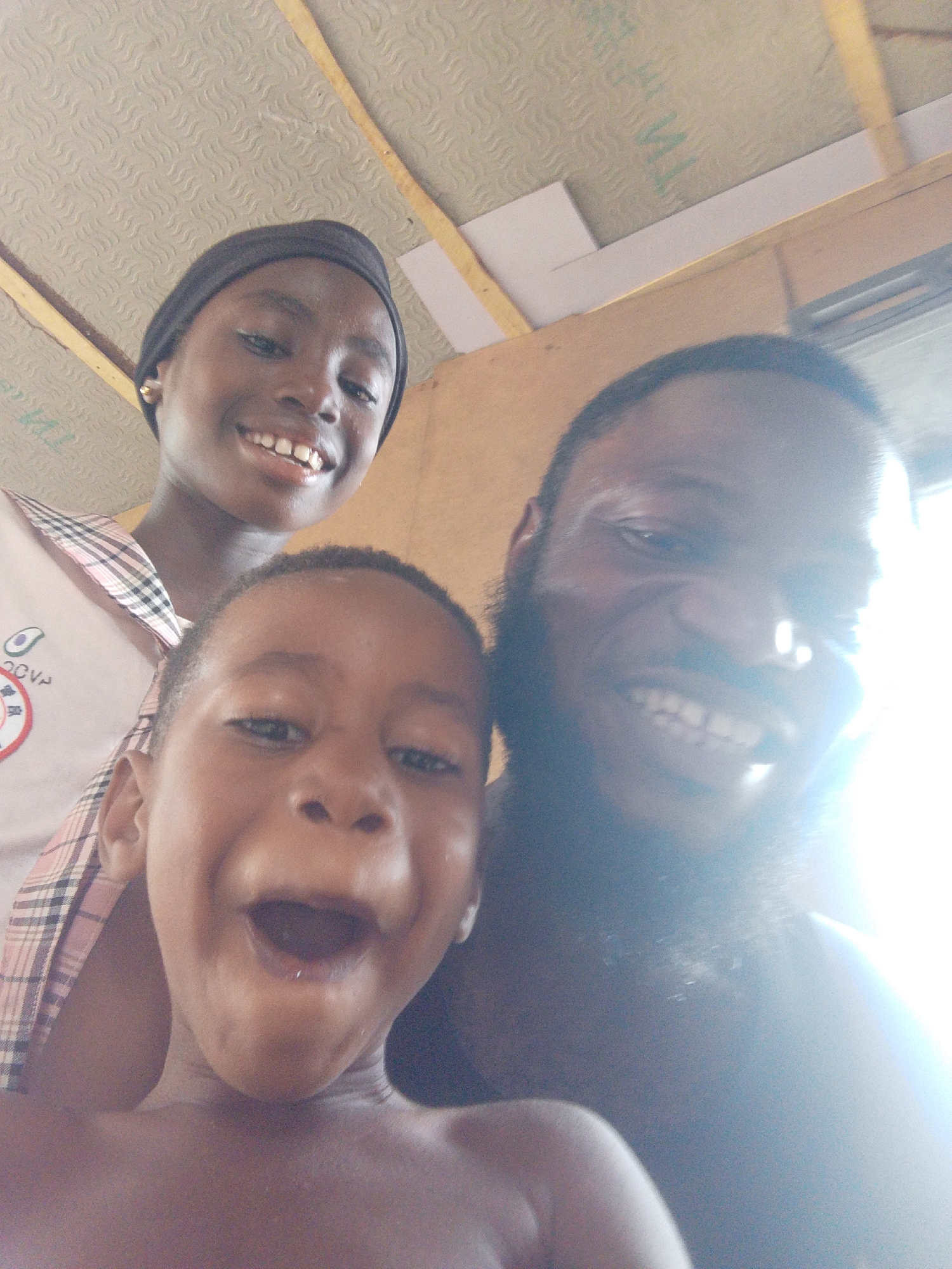 A close-up, high-angle selfie captures three individuals – a young girl, a young boy, and an adult male – smiling broadly, indicating joy and excitement. The setting is Lokoja, Nigeria.
The young girl, positioned in the upper left, wears a black head covering and a light-colored top with a pink and white plaid pattern on the collar/shoulder. A partially visible logo on her top shows what appears to be the letters "OVN" above an emblem. She grins, revealing her teeth.
The young boy is in the bottom center, with his mouth wide open, possibly laughing loudly or expressing surprise, and appears bare-chested.
The adult male, on the right, has a full beard and is also smiling broadly, though his face is partially obscured and overexposed by a bright light source coming from the right, creating a significant lens flare. He appears bare-chested or wearing a dark top.
The background suggests an indoor or covered space. A light-colored ceiling with a textured, possibly corrugated or patterned surface is visible at the top. Wooden beams or supports run across the ceiling. Faint, blurry green text, possibly "MA" or "WA," is barely discernible on a pale green section of the ceiling. The overall lighting is very bright, consistent with daytime, and contributes to the overexposure on the right side of the image. The atmosphere is clearly cheerful and lively.