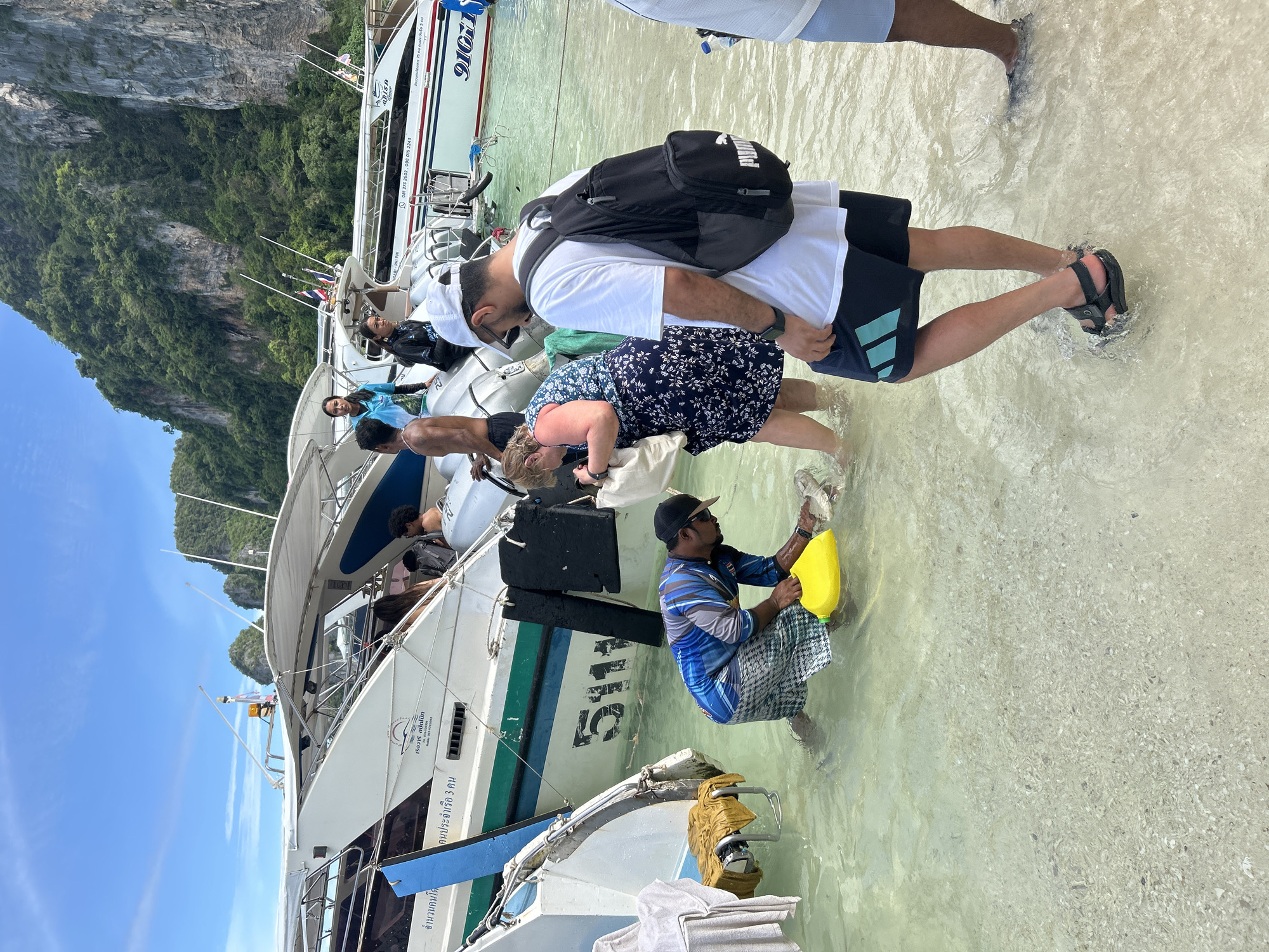 The image depicts a daytime scene on a shallow beach in Ao Nang, Thailand, with several speedboats engaged in passenger transfer. The water is clear, revealing a light-colored seabed of sand and shell fragments, with people wading in water reaching calf to knee height.
In the center-left, a man wearing a blue and black patterned long-sleeve shirt, checkered shorts, a dark baseball cap, and a watch is bent over in the water. He holds a bright yellow plastic container in his right hand and has his left hand submerged, interacting with a submerged object. To his right, a woman in a dark floral dress wades, carrying a light-colored bag over her left shoulder. Further to the right and slightly behind, a man with his back mostly to the camera is wading, wearing a white t-shirt, dark shorts with turquoise accents, black sandals, a white baseball cap, and a black Puma backpack. He also has a smartwatch on his left wrist. The lower legs of another person in light blue shorts are visible on the far right.
Multiple white speedboats are positioned in the shallow water. The closest boat on the left displays "5111" on its stern, along with blue and green stripes, and towels are draped over a rear railing. Another speedboat to the right features the text "KRABI - PHI PHI," partially visible numbers "910," Thai script, and phone numbers "081 273 2603, 096 015 2243." Several individuals are visible on the boats, some seated, some standing. Thai flags are flying from antennae or poles on the boat roofs.
The background is dominated by large, steep limestone karsts, densely covered in green tropical vegetation, under a bright blue sky with sparse white clouds.