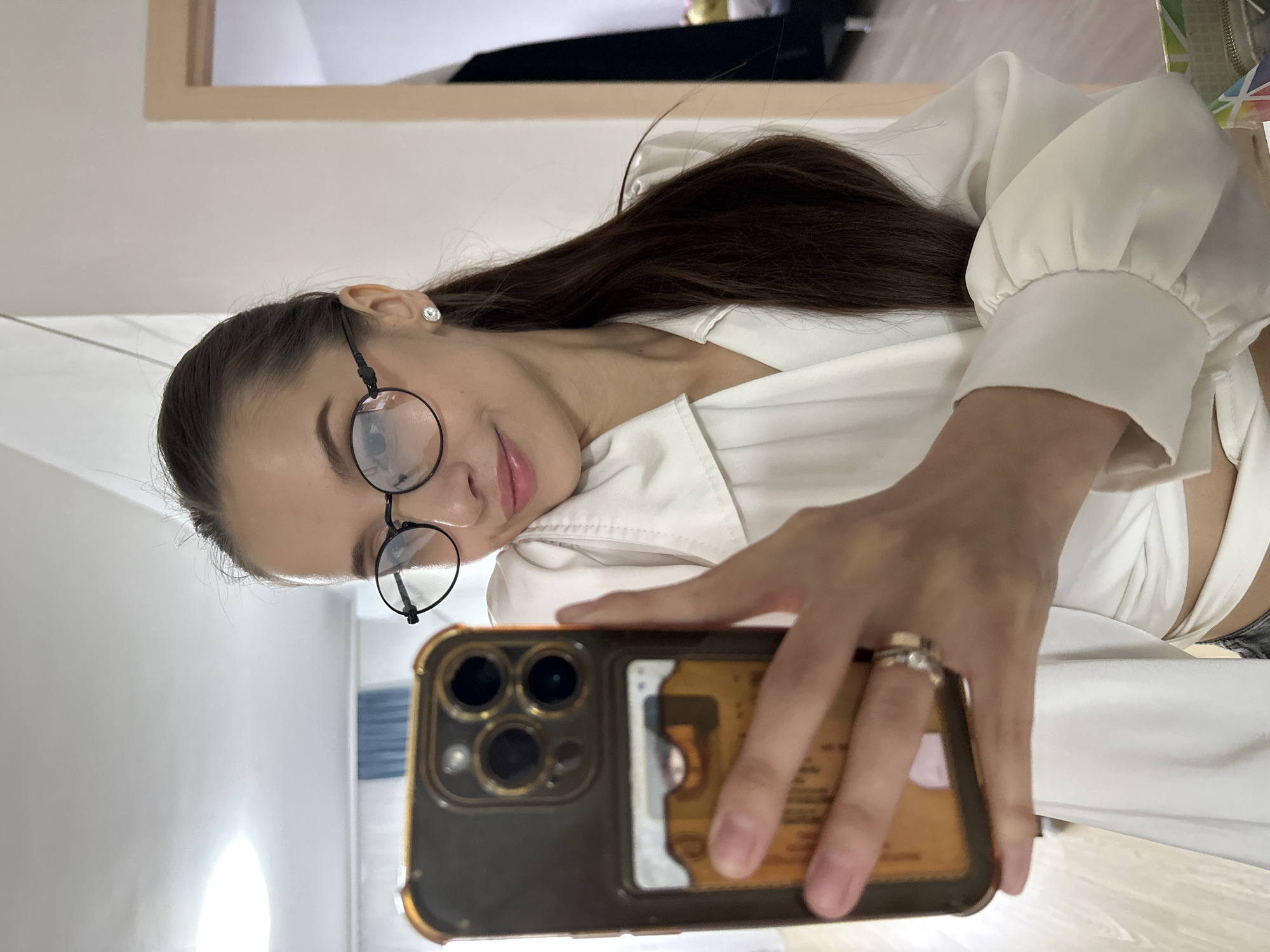The image captures a woman taking a mirror selfie, with her head tilted and a soft expression on her face as she looks towards the phone. Her long, dark hair is visible, falling over her shoulder. She wears round, dark-framed glasses and a single stud earring in her right ear. Her attire consists of a white, long-sleeved top with gathered cuffs, possibly a wrap-around style. A ring is visible on her right ring finger.
She holds a smartphone, identifiable by its triple-lens camera array on the back, in a dark case with gold-colored edges. In a transparent section on the phone's case, several cards are visible, appearing to be a credit card and an identification card, though no text is legible.
The background depicts a brightly lit indoor environment with light-colored walls. A rectangular mirror frame is partially visible in the upper background, confirming the selfie is taken through a reflection. The overall lighting is bright and even. The image is oriented with a significant counter-clockwise rotation. The scene suggests a casual moment indoors, likely in Yekaterinburg, Russia.