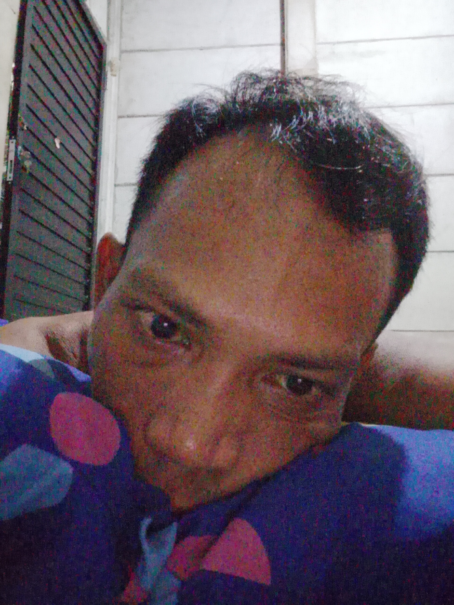 The image depicts a close-up selfie of a man with dark hair. He appears to be lying down, with his face close to a patterned blue and pink pillow. The background shows a door with dark slats and a white wall. The photograph was likely taken indoors. The location is Bukit Ketok, Indonesia. No specific activities, interactions, or other subjects or objects are discernible.