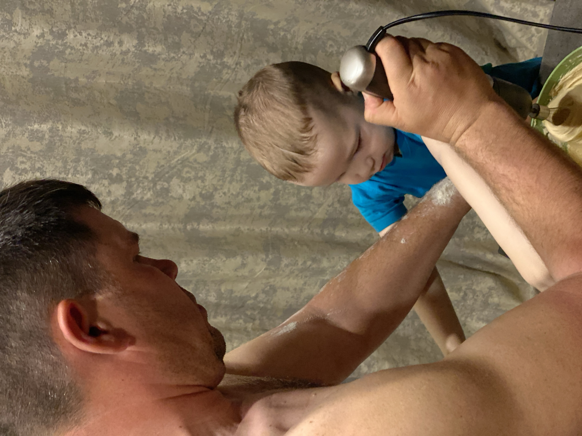 A man and a young child are engaged in baking. The man is shirtless, with a dusting of flour on his arm. He is holding an electric mixer over a green bowl containing batter. The child, wearing a blue shirt, is standing close to the man, watching the mixer. The background is a patterned surface. The location is not discernible.
