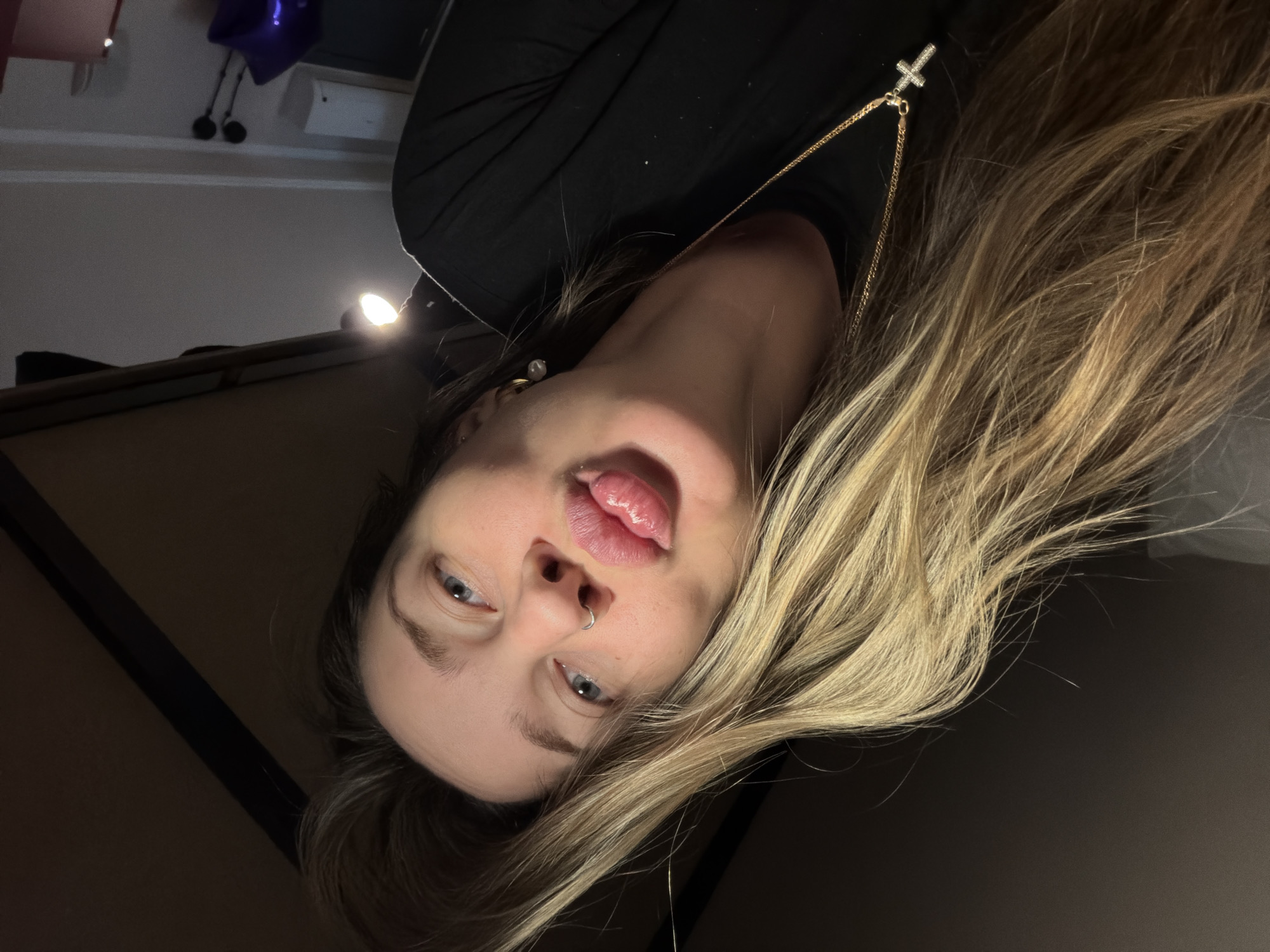 A young woman with long, wavy blonde hair is depicted in a close-up, head-and-shoulders shot, oriented upside down. She is looking directly at the camera with a playful expression, slightly sticking out her tongue. Her eyes are a striking blue. A small silver hoop nose ring is visible on her right nostril. She wears a black top or sweater, and a gold chain necklace with a small, diamond-encrusted cross pendant rests on her chest. A pearl earring can be seen on her left ear.
The scene is dimly lit, suggesting an indoor setting during evening or night. A bright, localized light source is visible in the upper right portion of the frame (when viewed in its original upside-down orientation), casting soft illumination on her face and hair. The background is dark and largely indistinct, with faint outlines of what appears to be a wall or ceiling structure. The overall impression is casual and intimate. No visible text is present in the image itself. The provided location context is Moscow, Russia.