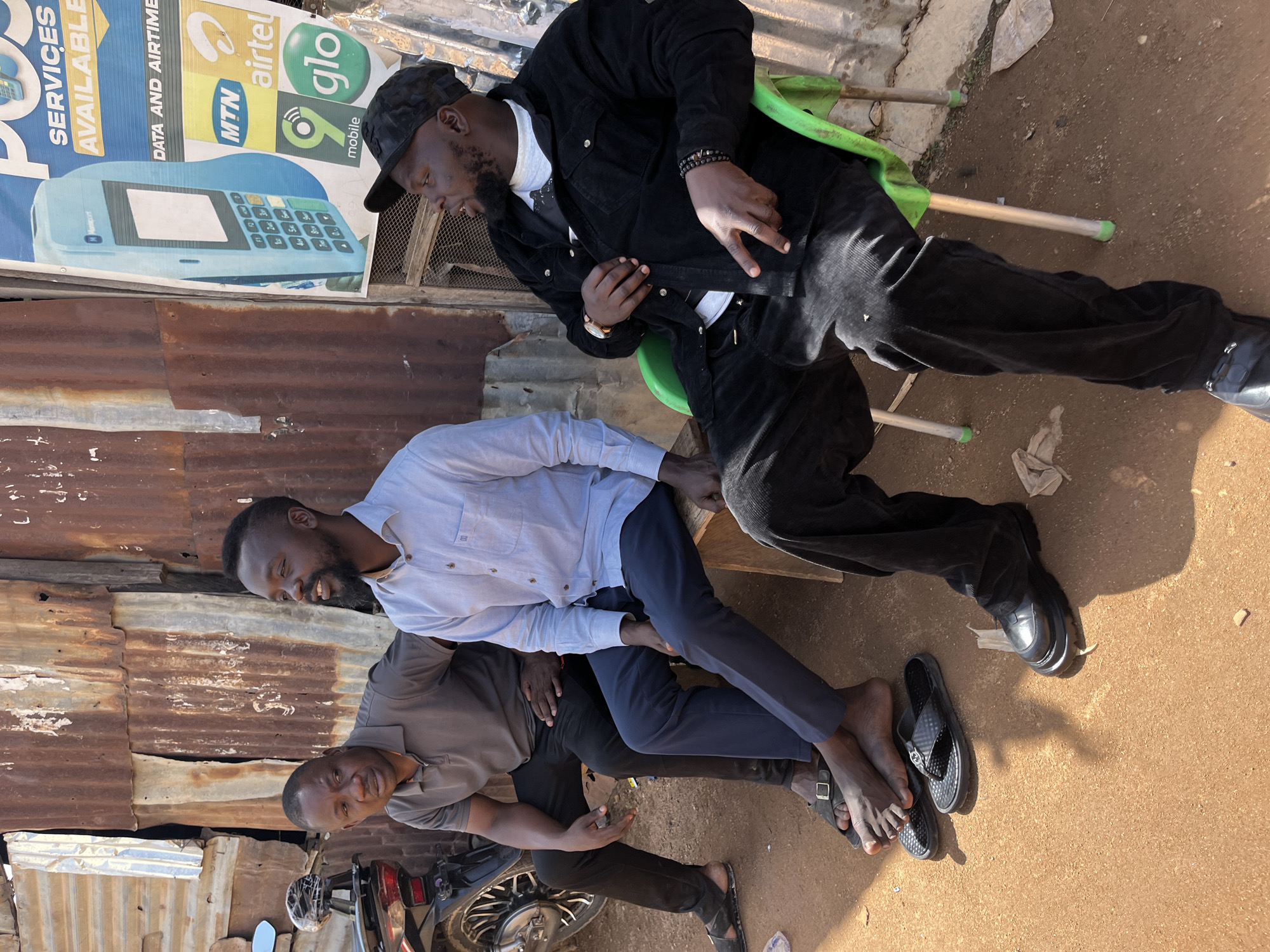 This image captures three men socializing outdoors in Jos, Nigeria, on a bright, sunny day, evidenced by the strong shadows. The setting features a rustic backdrop of rusted corrugated metal sheets, with a dirt ground beneath.
On the right, a man with a beard and a black cap is seated on a green plastic chair. He is dressed in a black jacket, white t-shirt, black pants, and black shoes, accessorized with a watch and beaded bracelets. He looks down or to his side.
In the center and left, two other men are seated on a low wooden bench or platform. The man in the center, also with a beard, wears a light blue long-sleeved shirt and dark pants, and is smiling towards the third man. The third man wears a grey polo shirt and black pants; his black sandals are on the ground beside his bare feet. He looks towards the camera with a subtle smile. Parts of a motorcycle are visible in the lower left corner of the frame.
A large banner behind the men advertises various mobile services. Clearly visible text includes "SERVICES AVAILABLE" and "DATA AND AIRTIME," along with the distinct logos for "MTN," "airtel," and "glo mobile." The banner also displays an image of a light blue Point-of-Sale (POS) device, with "Newport" visible on its front. The scene suggests a casual gathering in a local business or public area.