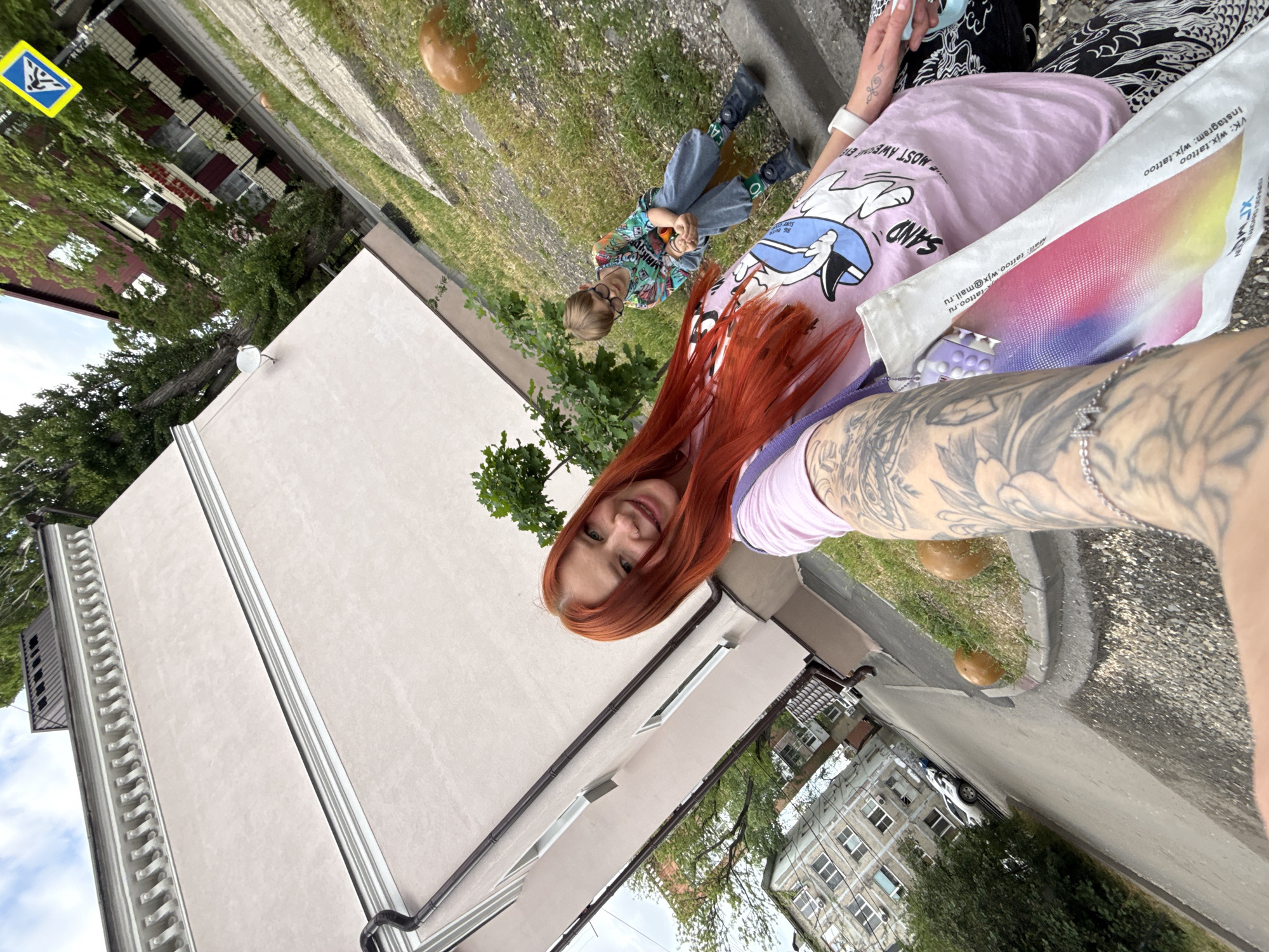 A woman with red hair and visible tattoos on her arm is taking a selfie. She is wearing a pink shirt with a cartoon graphic. A child, possibly a boy, wearing glasses and a colorful patterned shirt is sitting on the ground. A shopping bag is next to the woman. The background includes buildings, grass, and a street. The OCR in the image mentions "wjx.tattoo," suggesting a connection to tattoos, Rostov-on-Don, Russia. The sign suggests a possible location in Russia.