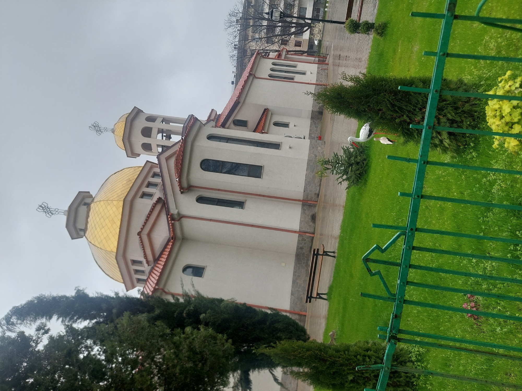 The image shows the Church of the Holy Mother of God in Asenovgrad, Bulgaria. The church has a white facade with a red roof and a golden dome. A green fence is in the foreground, and there are trees and grass around the church. There is a bench near the church. A pair of decorative storks stand in the garden. The sky is overcast.