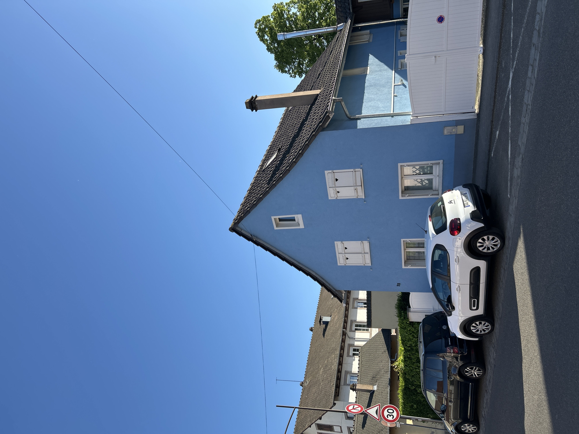 This image captures a bright, sunny street scene in a residential area, likely in Strasbourg, France, as indicated by the context. The dominant feature is a vibrant blue two-story house with a dark tiled roof.
The blue house features white-framed windows, some equipped with white shutters, two of which have decorative heart-shaped cutouts. A concrete chimney stack and a modern metallic flue extend from the roof. The house number "32" is visible on the blue facade. To the right of the main house, a white garage door or gate is partly visible.
Two cars are parked on the asphalt road in front of the blue house. Closest to the viewer is a white Citroën C3 Aircross, with a partially visible French license plate that reads "AA-716-CG". Behind it, a dark-colored SUV, possibly a Volkswagen, is also parked.
The sky is a clear and intense blue, indicating bright daylight and good weather. A thin, dark wire or cable stretches diagonally across the sky. Green foliage from trees and hedges can be seen in the background and between buildings. Other residential houses with dark tiled roofs and lighter facades line the street further back.
On the left side of the street, two road signs are visible: a circular speed limit sign displaying "30" (km/h) and another partially obscured circular sign with a red border. Shadows are cast by the cars and buildings, suggesting midday to early afternoon.