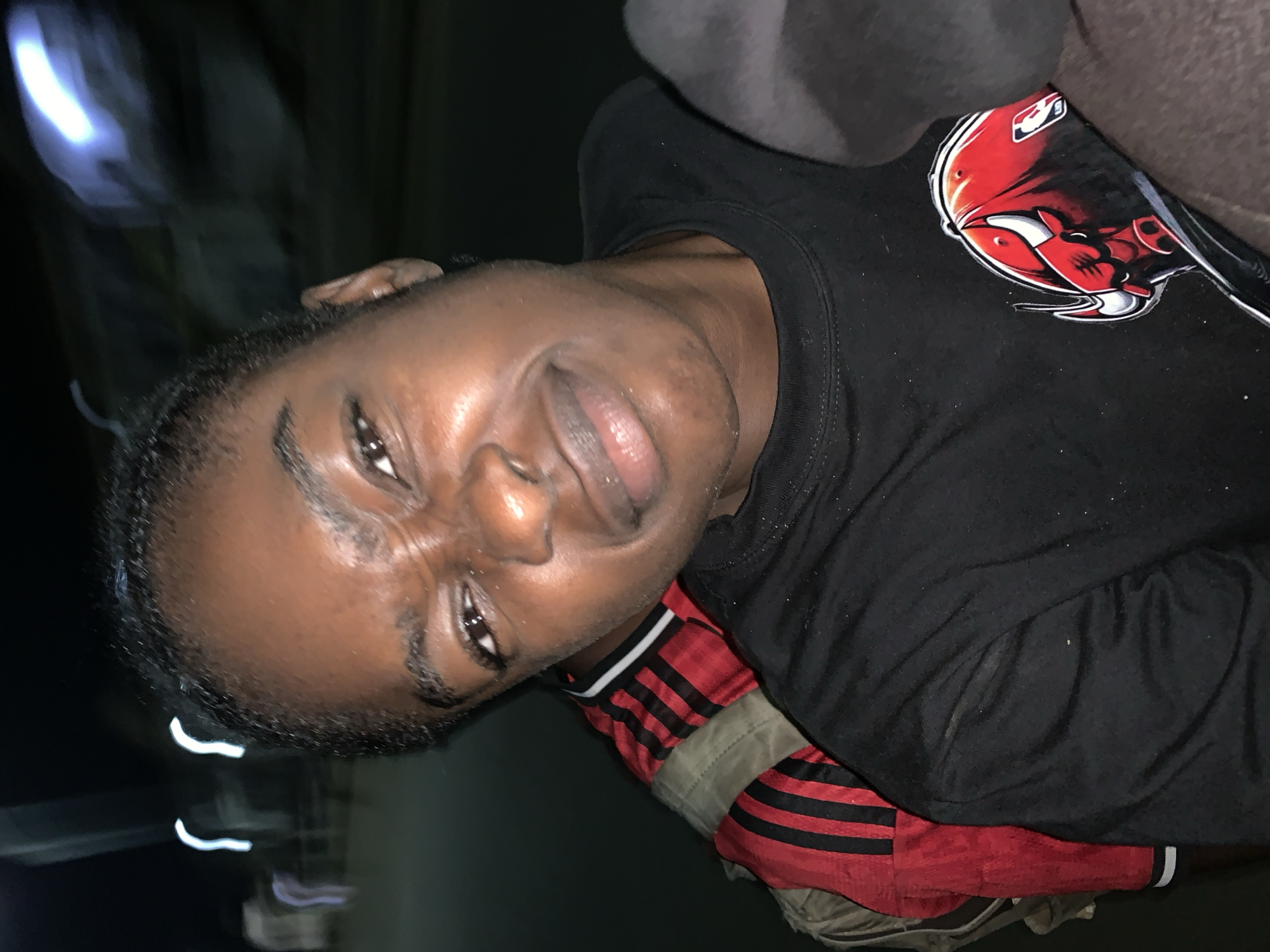 A dark-skinned person wearing a black long-sleeved shirt with a Chicago Bulls logo is visible from the chest up. Underneath the black shirt, a red garment with black stripes is partially visible. The person is indoors, and a blurry object appears on the left side of the frame. There is a gray cushion or headrest in the background. The image is rotated counter-clockwise. The city of Kabong, Nigeria, cannot be inferred from the image.