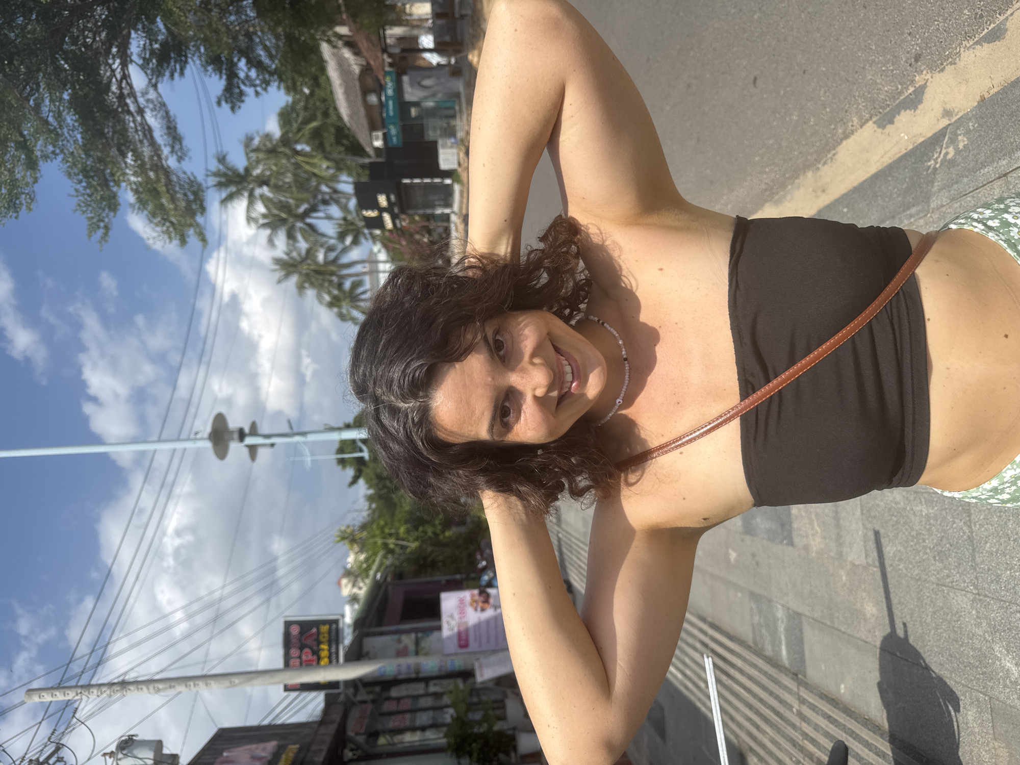 A young woman with dark, curly hair is posing for a picture outdoors on a sunny day. She is wearing a black strapless top and a green and white patterned skirt. Her arms are raised, and she is holding her hands behind her head, smiling at the camera. A thin brown strap from a purse or bag is visible across her chest.
She is standing on a paved sidewalk next to a street. In the background, there are buildings with signs, palm trees, and a blue sky with white clouds. The scene appears to be in a tropical or subtropical urban environment. Based on the presence of palm trees and the style of architecture, it is likely a tourist area. The sign in the background, partially visible, reads "MASSAGE" with some Vietnamese text. This suggests a commercial area that might cater to tourists, such as a street in Mũi Né, Vietnam, known for its beaches and resorts. The overall atmosphere is casual and relaxed, with the bright sunlight indicating daytime.
