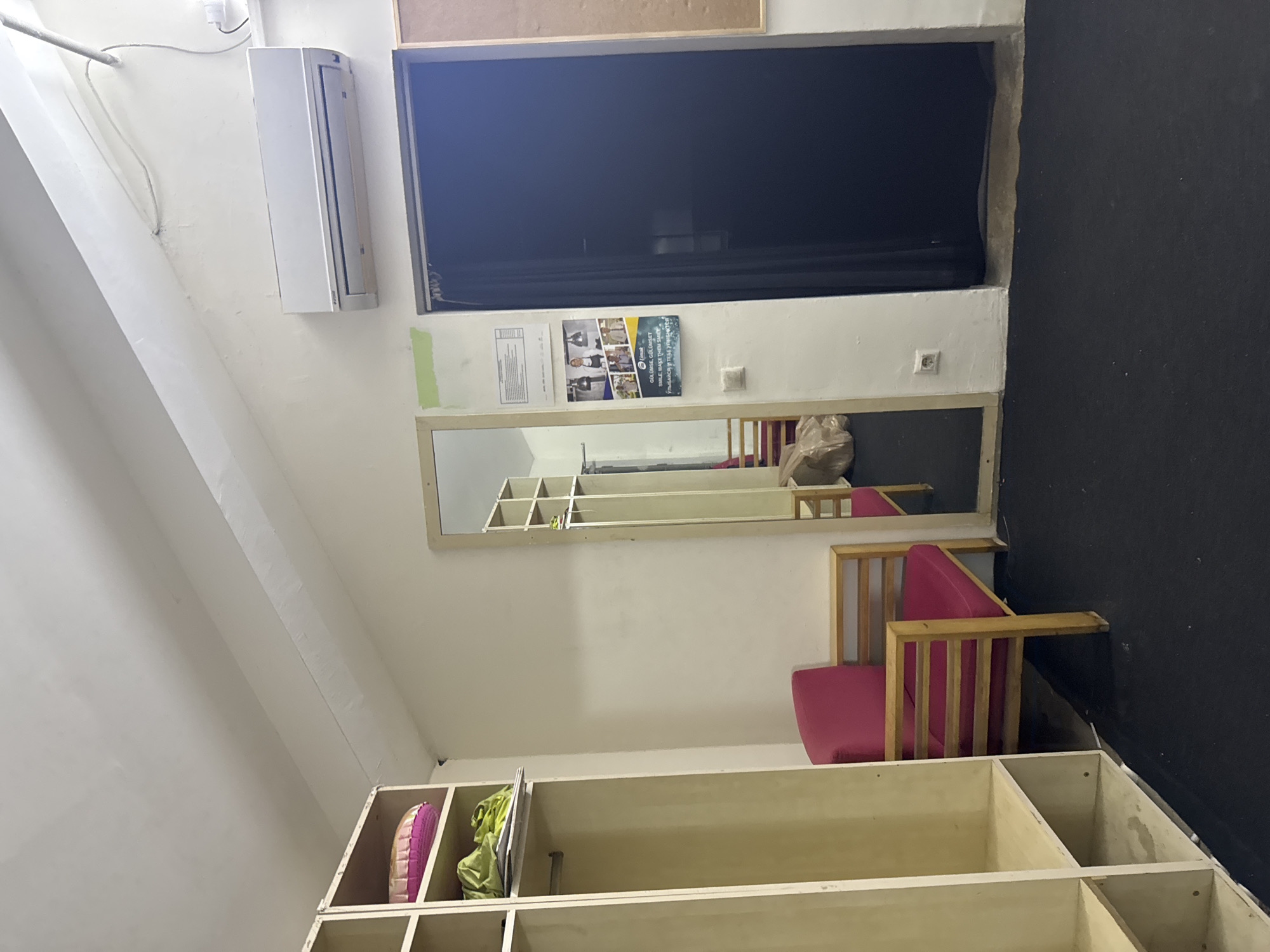 An interior scene captures a dimly lit room that appears to be a changing or waiting area. The room is furnished with a tall, open shelving unit on the left, filled with various items including a round pink cushion and green fabric. To the right of the shelving unit is a bright pink upholstered chair with a light-colored wooden frame. A full-length mirror leans against the wall further to the right, reflecting another similar pink chair and a stack of brown paper bags. Adjacent to the mirror is a closed dark blue curtained doorway. Mounted high on the wall above the doorway is a white air conditioning unit. On the wall to the left of the doorway, a poster with images of people and text in what appears to be Turkish and Cyrillic script is displayed, along with a smaller document above it. The floor is covered in a dark, textured material, and the walls are painted a pale off-white. The ceiling slopes upwards, with exposed beams or structural elements visible. The overall impression is of a functional, somewhat spartan space, possibly in a fitness center, performance venue, or similar establishment. The image location is stated to be in Aksu, Turkey.