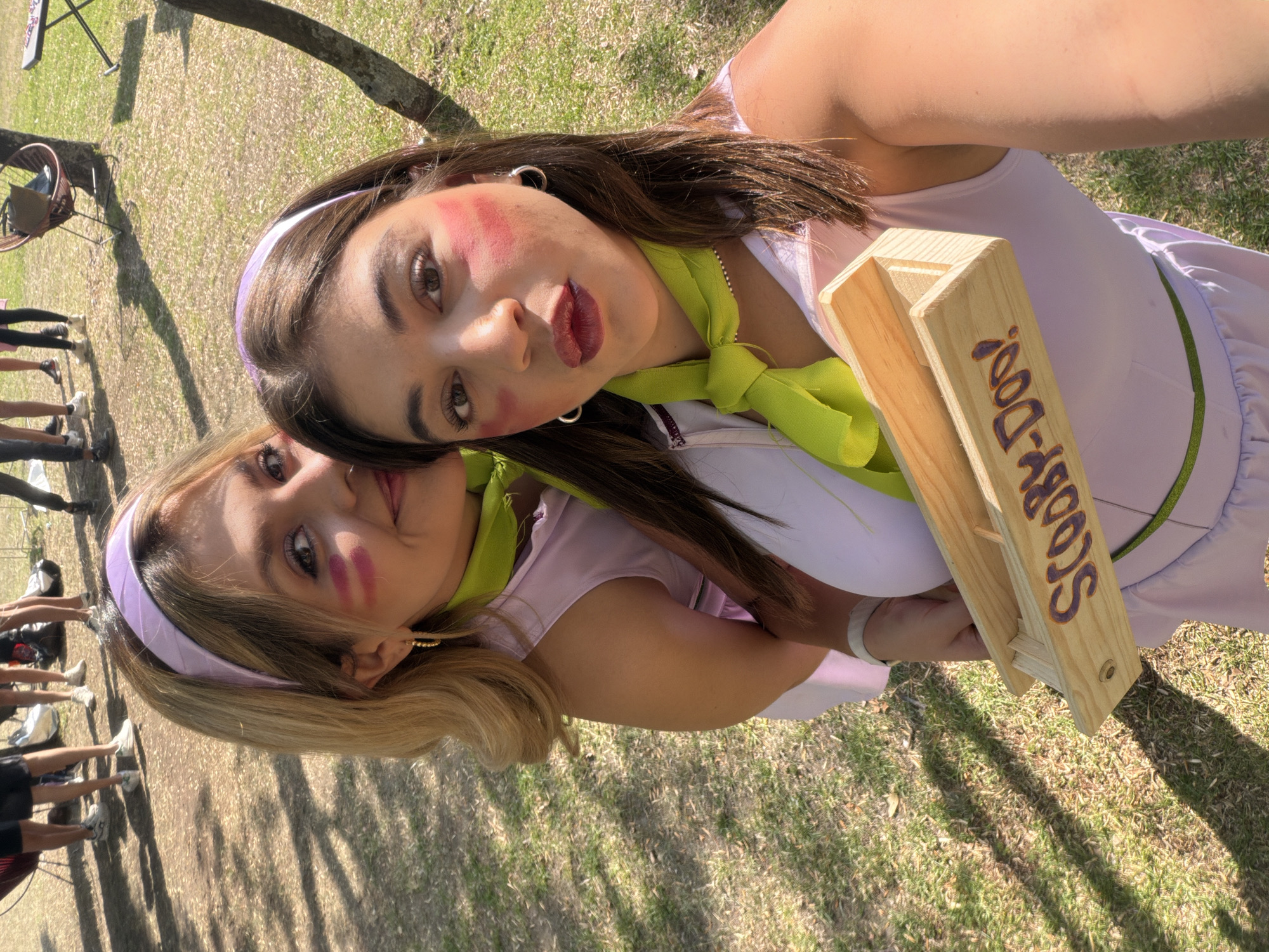 Two women are in the foreground of an outdoor photo, likely a selfie. Both women are wearing light purple headbands and light green neck scarves. The woman in the upper right has dark brown hair and is wearing dark red lipstick with pink blush on her cheeks. The woman in the lower left has blonde hair and pink blush on her cheeks. They are holding a wooden object with the words "Scooby-Doo" written on it in purple. The background shows green grass and trees with other people in the distance. The location is San Bernardino Tlaxcalancingo, Mexico.