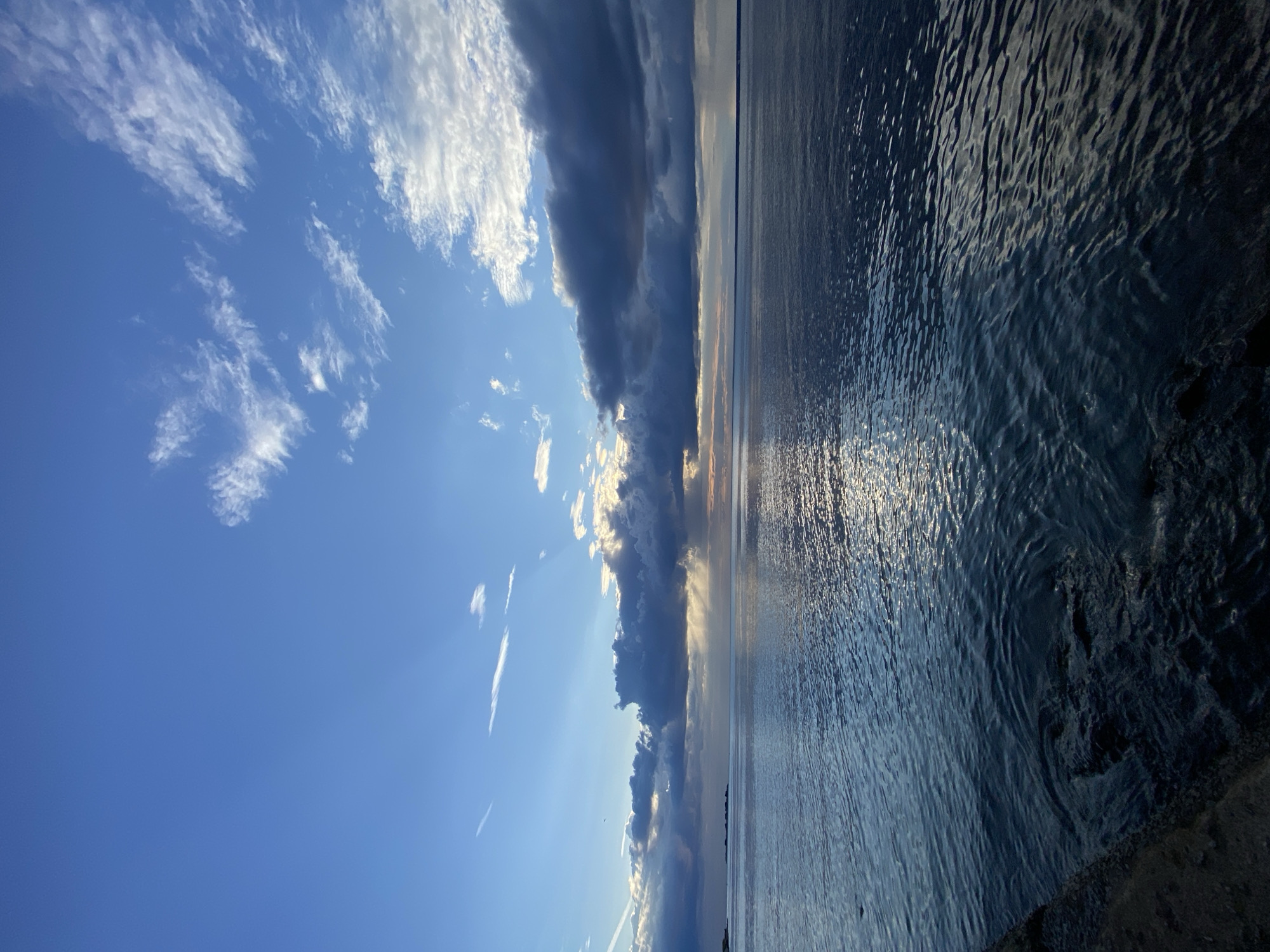 A wide-angle, vertical outdoor photograph captures a view of the Gulf of Finland from the Sestroretsk embankment in Sestroretsk, Russia. The scene is dominated by a vast expanse of dark blue water in the foreground, reflecting the sky. The horizon line is visible in the distance, with a faint landmass or islands on the far left. Above the water, the sky is a gradient of deep blue to lighter blue, with scattered clouds. Dark, heavy clouds are clustered in the middle of the frame, with sunlight breaking through them in visible rays, creating a dramatic effect. The water is slightly rippled, suggesting a gentle breeze. The lower left of the frame shows a dark, textured surface which appears to be the edge of the embankment or a seawall. No people or animals are visible in the frame. The lighting suggests it is late afternoon or early evening.