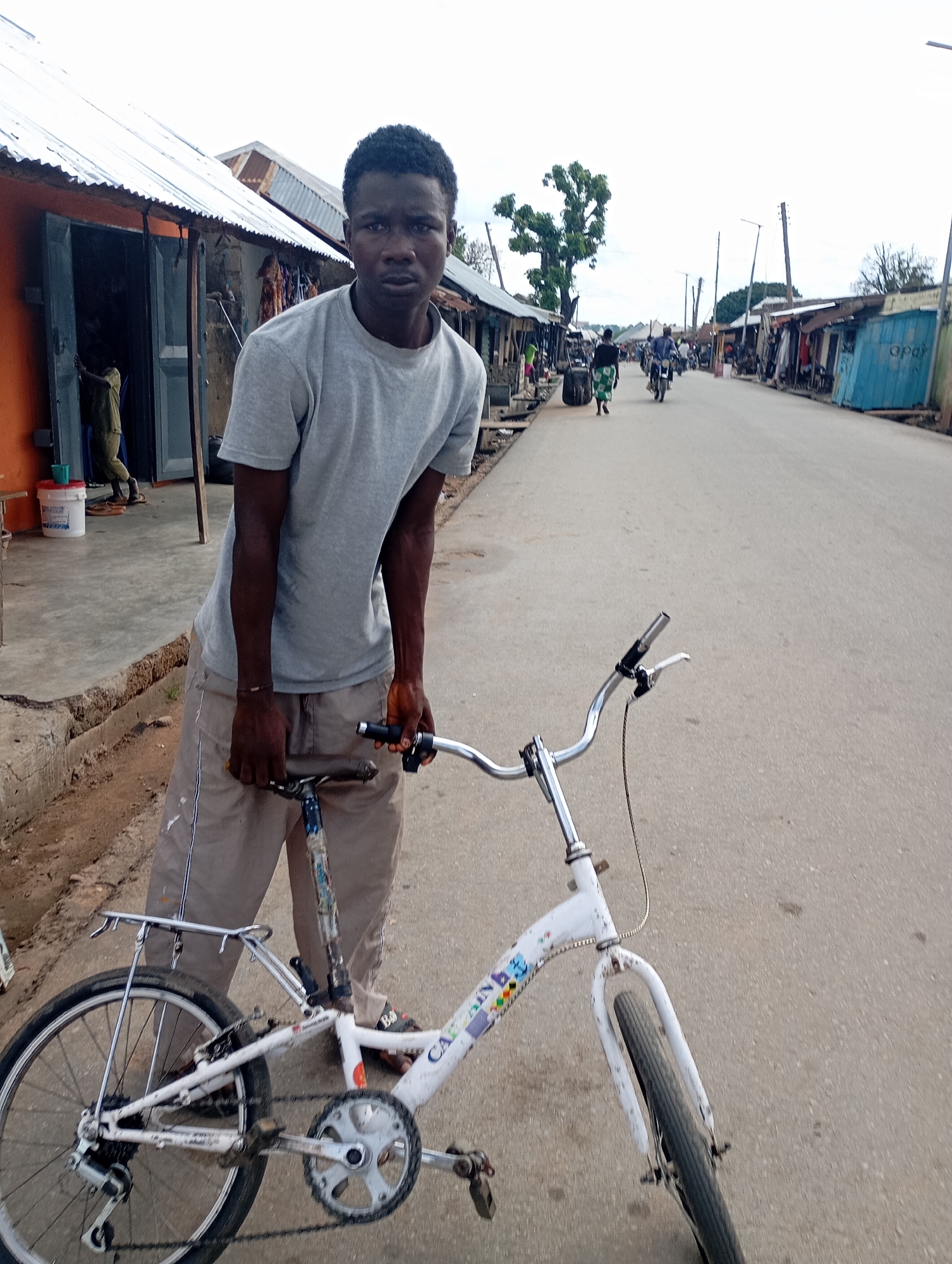 A young Black male stands with a white bicycle on a paved street in the city of Shendam, Nigeria. He is wearing a grey t-shirt and tan pants. The bicycle is a road bike with a rear rack. In the background, there are buildings with corrugated metal roofs and various people walking and riding motorcycles. The sky is overcast. The scene appears to be a busy market street.