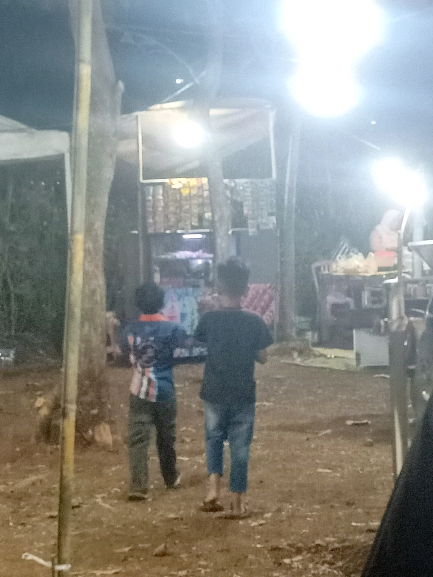 The image depicts an outdoor scene at night, possibly a market or a gathering, in the city of Tasikhargo, Indonesia. Two young boys, viewed from behind, are walking away from the camera. The boy on the left is wearing a dark long-sleeved shirt with a graphic design and dark pants. The boy on the right is wearing a dark short-sleeved shirt and blue jeans. They are walking on a dirt ground littered with debris. In the background, there are stalls or structures with goods displayed. Bright lights illuminate the scene, creating glare and making some details indistinct. There appears to be vegetation, including trees, on the left side of the image. The overall atmosphere suggests a lively but somewhat chaotic environment.