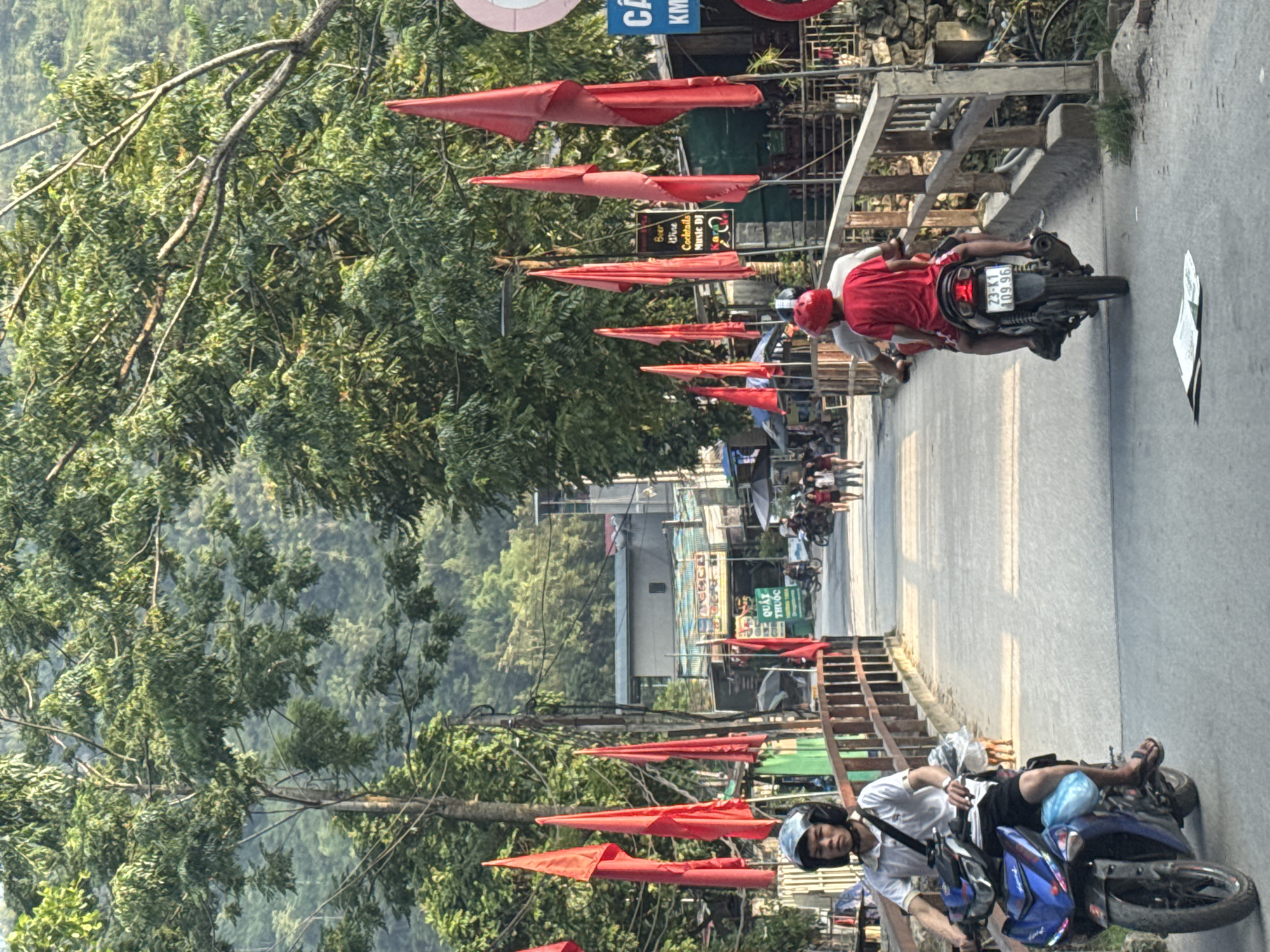 The image captures a road scene in Du Già Commune, Vietnam. The road is adorned with a series of red, cone-shaped flags suspended overhead. Two motorbikes are visible on the road. One has two male riders, the driver wearing a red shirt and helmet. The motorbike's license plate reads "23-K1 109.96." The second motorbike has a single male rider, wearing a white shirt and blue helmet. The background features lush green trees and mountains. The architecture of buildings are visible along the road.