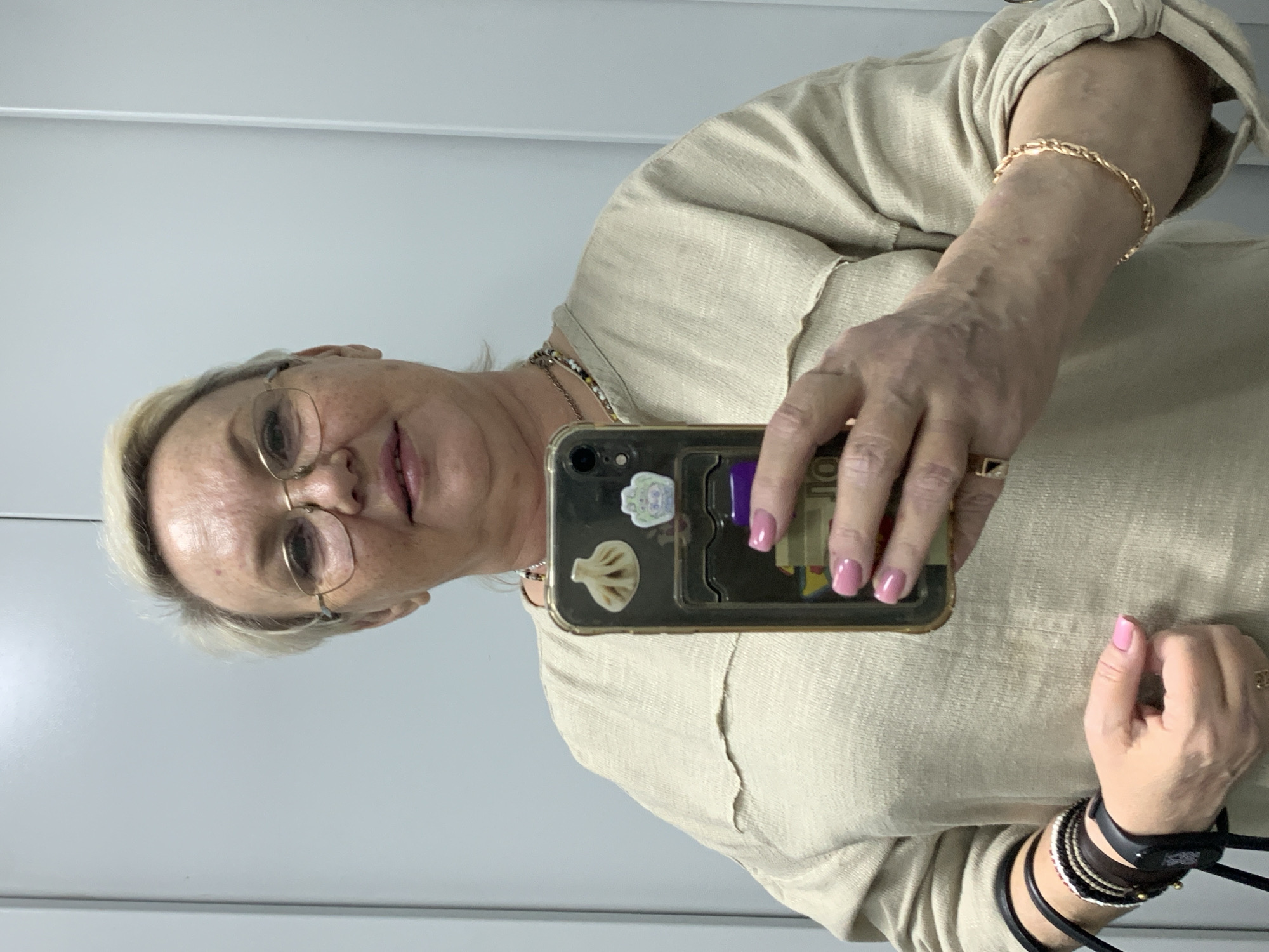 A fair-skinned woman with short blonde hair and glasses is holding a smartphone up to take a selfie in what appears to be an interior location with a white wall in the background. She is wearing a beige shirt or blouse, and various pieces of jewelry including a gold bracelet and several bracelets on her right wrist. The smartphone case has stickers on it, including a dumpling. Her fingernails are painted pink. The location is Chaikovsky, Russia.