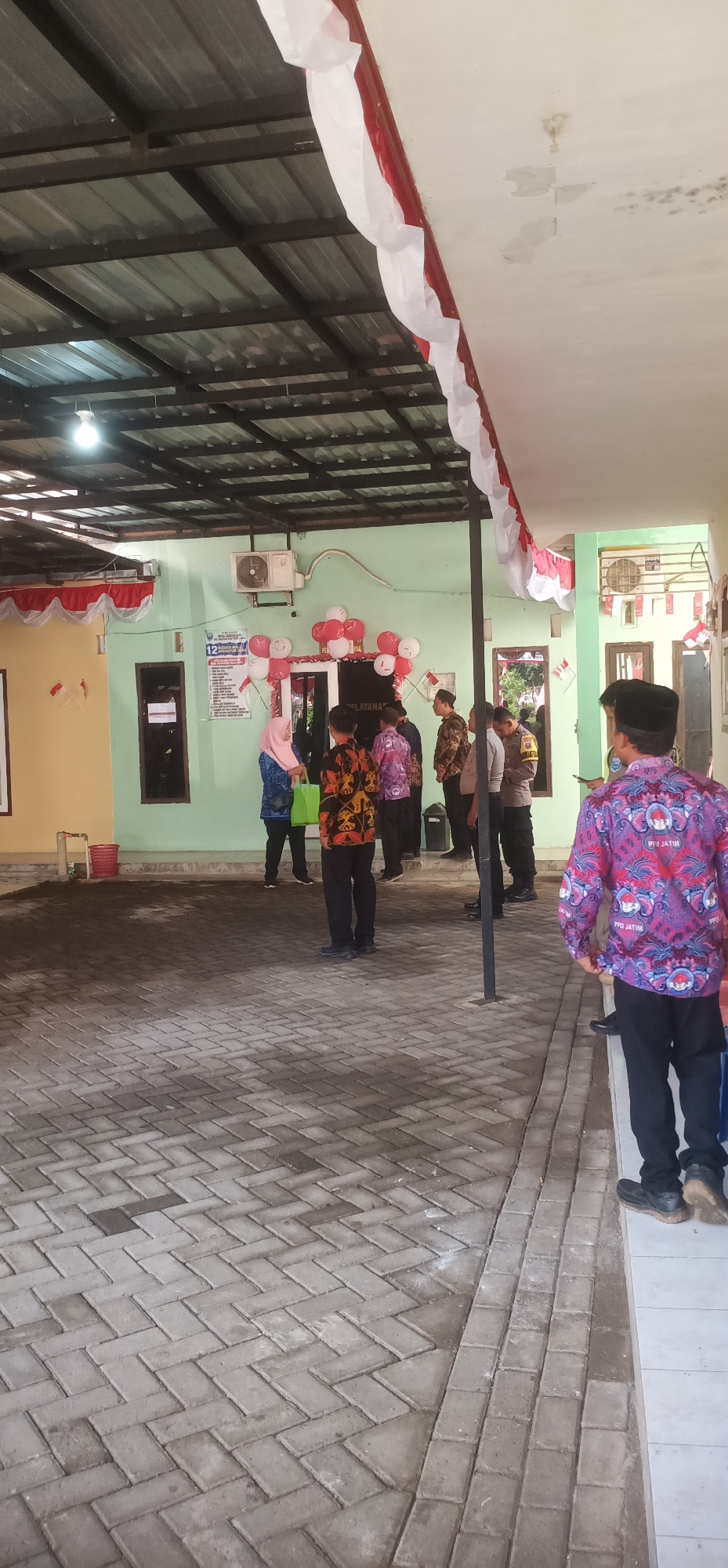 This image depicts an outdoor area in front of a building, likely in Tamansari, Indonesia, adorned with red and white Indonesian flags and banners. Several individuals are present, some appearing to be officials or employees, and others possibly visitors.
In the foreground, the ground is paved with interlocking concrete bricks arranged in a herringbone pattern. To the right, a person wearing a patterned purple shirt and black pants stands facing away from the camera.
The midground shows a light green building with several doors and windows. Red and white balloons are arranged in clusters around one of the doorways, and more flags are visible on the walls. A sign with Indonesian text is mounted to the left of the entrance. Several people are gathered in front of the building, including a woman in a pink headscarf, individuals in patterned shirts, and people in what appear to be official uniforms. An air conditioning unit is mounted on the building's exterior.
The background reveals a covered outdoor area with a corrugated metal roof supported by dark metal beams. A light bulb is visible hanging from the ceiling. Further to the right, another section of the building is visible, also decorated with Indonesian flags. The overall scene suggests an event or a gathering taking place at this location.