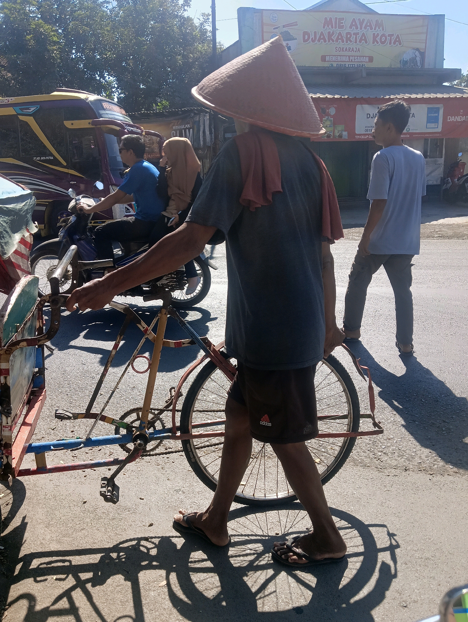 A man wearing a conical hat walks a pedicab on a sunny day in Sokaraja, Indonesia. The pedicab, which appears to be made of a bicycle frame and a carriage, is partially visible on the left side of the image. The man is holding onto the handlebars of the pedicab, with his right hand on the frame. He is wearing a dark grey t-shirt, dark shorts, and flip-flops. A reddish-orange cloth is draped over his shoulders. The pedicab's wheels are visible, as are the pedals and chain. In the background, a motorcycle with two riders, a man and a woman, is stopped. The woman is wearing a hijab. A bus is also visible behind the motorcycle. A building with a sign that reads "MIE AYAM DJAKARTA KOTA" is in the background. The street is paved and appears to be in a commercial area. The scene is illuminated by bright sunlight, casting shadows on the ground.