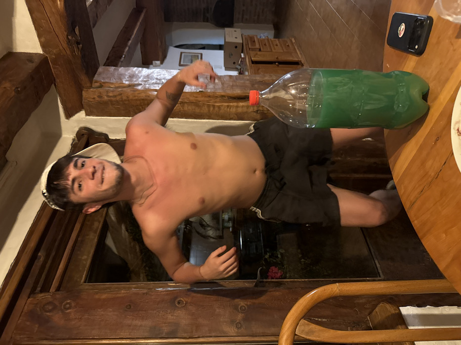 A shirtless man wearing a hat and black shorts poses on a wooden structure. A plastic bottle filled with green liquid and a black phone case sit on a wooden table beside him. Wooden beams and furniture are visible in the background.