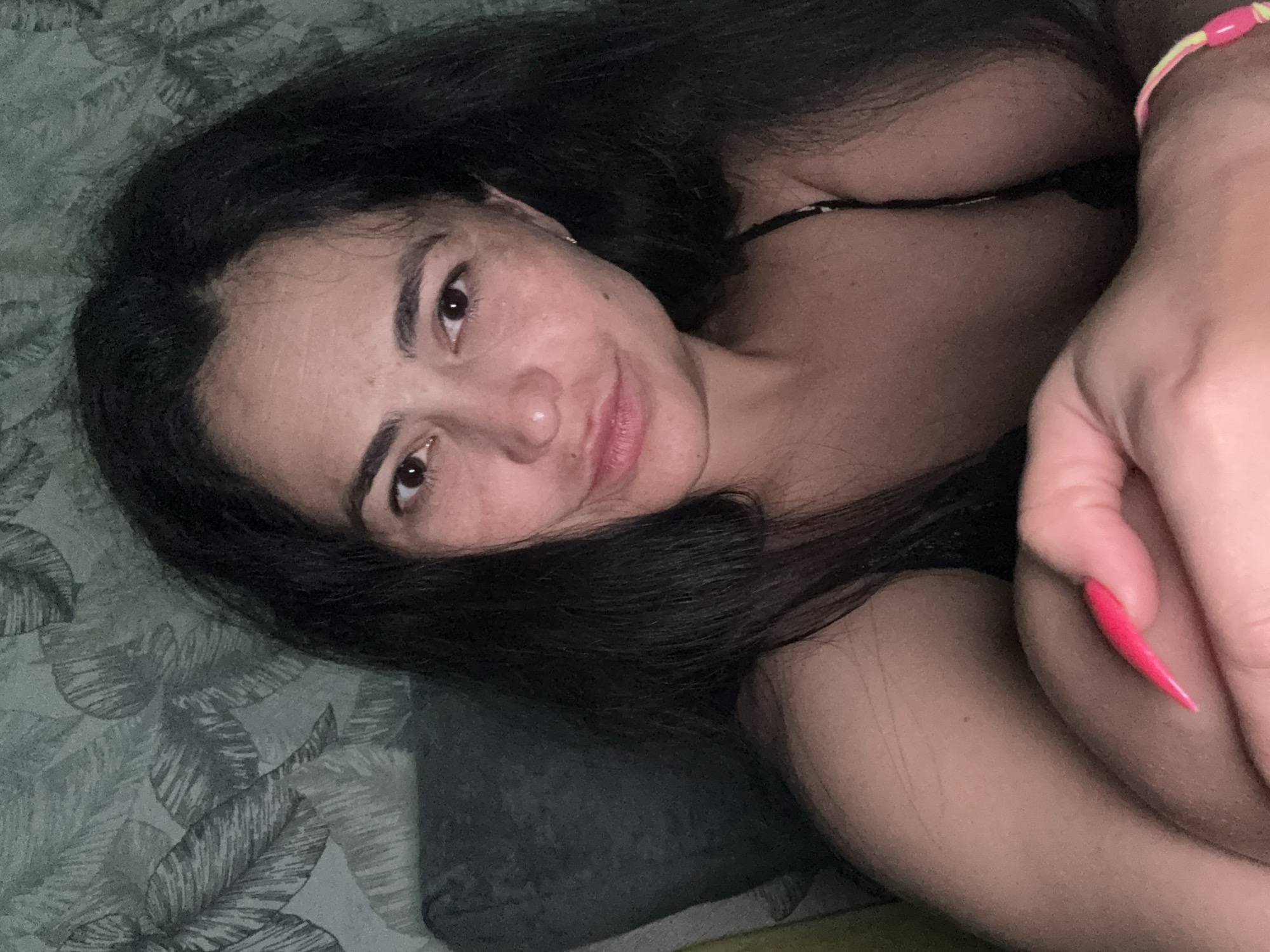A person with long dark hair is lying on a bed with a patterned bedspread. The person is facing the camera and smiling slightly. They have brown eyes and fair skin. Their right hand is raised near their face and a pink bracelet is visible on their wrist. The person has long pink nails. The location in the city of Kauchuk Stantsiya, Russia is unknown.