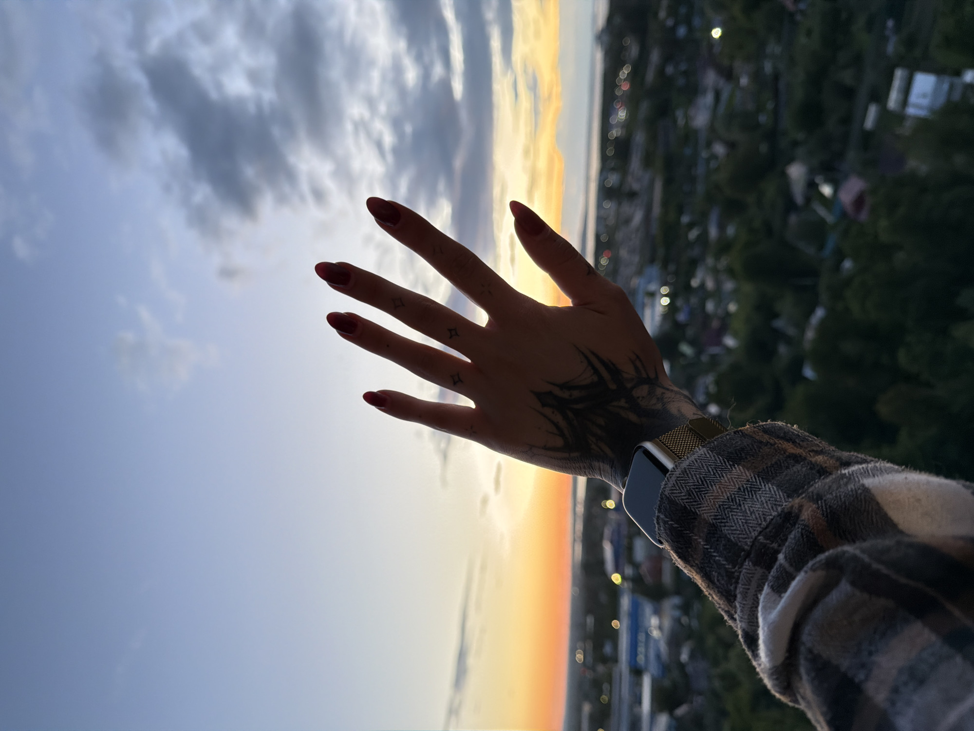 The image shows a person's hand reaching out towards a sunset. The hand has dark tattoos on the wrist and fingers, and the fingernails are painted red. The person is wearing a plaid shirt and a smartwatch with a gold band. The background is a blurred cityscape with trees and lights, suggesting an urban or suburban setting. The sky is a mix of clouds and a vibrant orange and yellow sunset, indicating it is evening. The composition is a close-up on the hand and arm, with the landscape and sky providing context. The overall mood is contemplative or peaceful, with a focus on the beauty of the sunset. There is no visible text in the image.