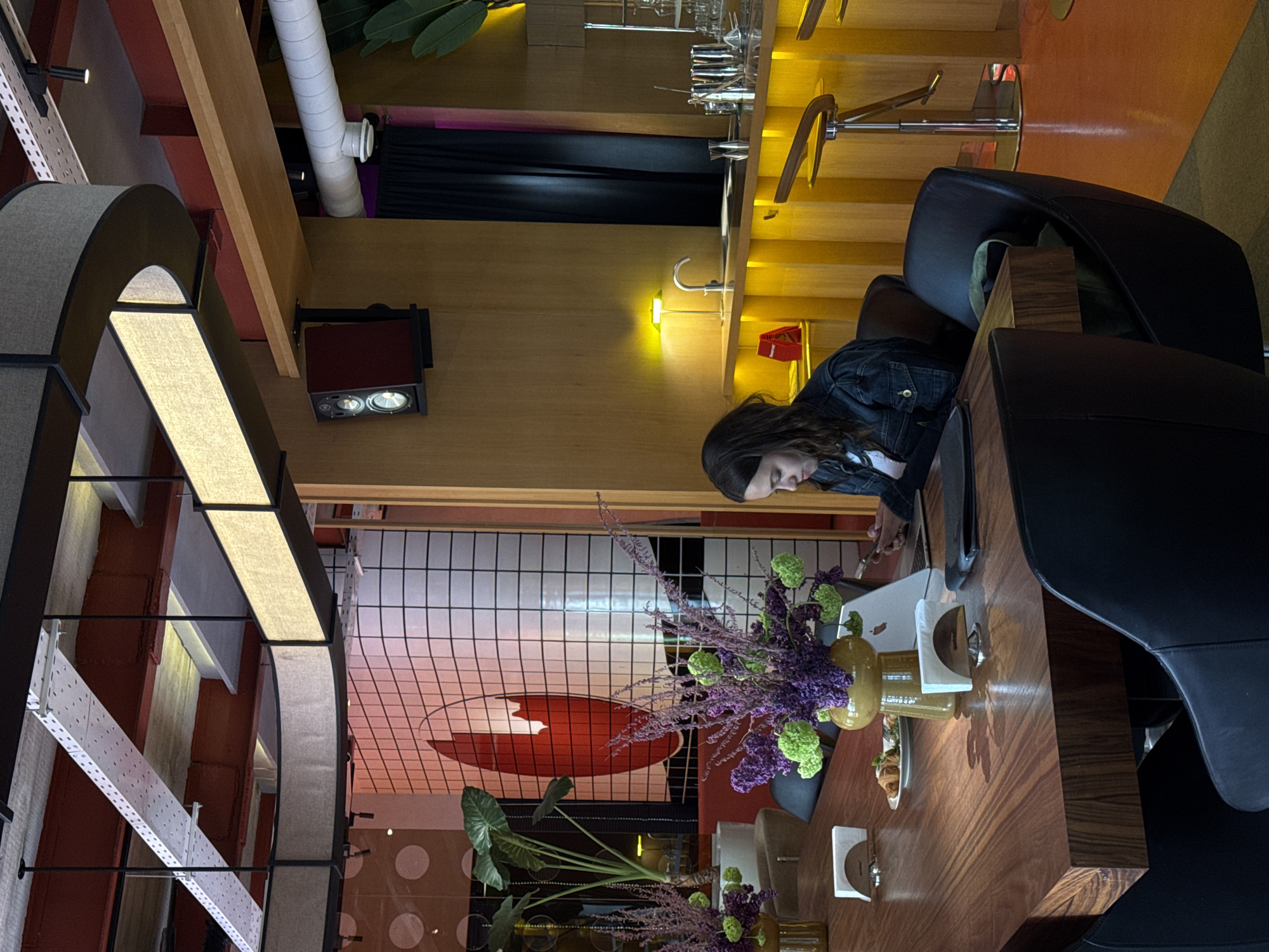 This image captures a modern and stylish interior space, likely a cafe, restaurant, or co-working area, located in Yerevan, Armenia. The scene is dominated by warm artificial lighting, suggesting an indoor setting irrespective of the time of day.
In the foreground and slightly to the right, a woman with dark hair, dressed in a denim jacket over a white top, is seated at a long, dark wooden table. She is looking down, seemingly focused on a laptop or tablet positioned in front of her. Her posture indicates engagement with her device.
The table features decorative elements, including a tall vase filled with striking purple and green floral arrangements, flanked by smaller glass bottles or vases. Black chairs with wooden backs are positioned around the table.
The interior architecture is characterized by a blend of materials and lighting. Light wooden panels adorn the walls, interspersed with exposed red structural beams and a white perforated metal railing. A large, arch-shaped ceiling light, constructed with fabric panels, provides diffuse illumination. Additional lighting comes from recessed spotlights, a wall-mounted fixture with two circular bulbs, and smaller yellow wall lamps, which cast a warm glow across the wooden surfaces.
A prominent feature in the background is a section of wall covered in white tiles with a black grid pattern, on which a large, abstract red design or logo is displayed. Potted plants with large green leaves are strategically placed, adding natural accents to the contemporary decor. Further into the background, a bar or counter area is visible, equipped with various metal fixtures and glassware. The overall ambiance is chic and inviting. No discernible text is visible within the image.