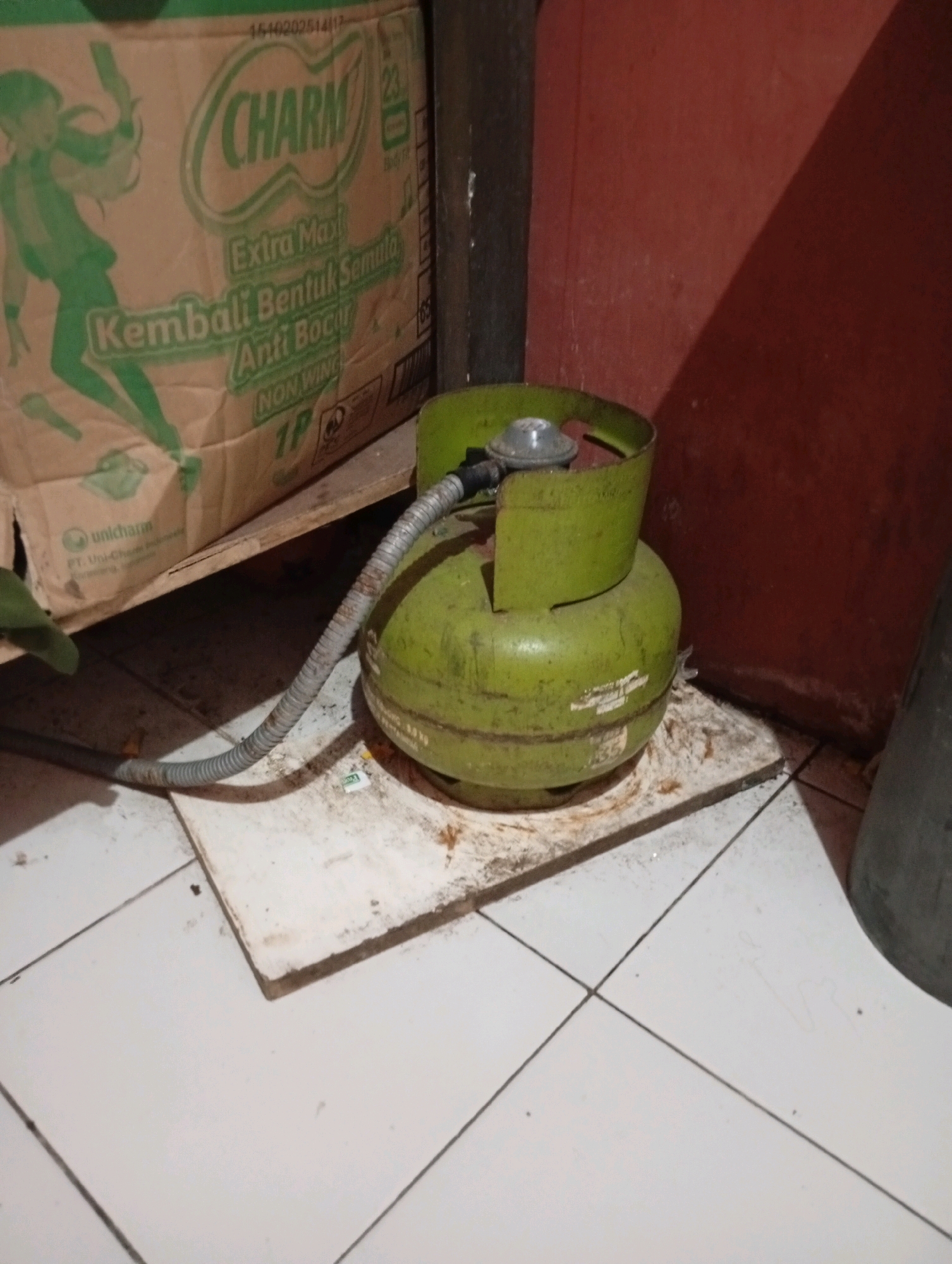 In an indoor setting, possibly a kitchen or storage area, a green LPG cylinder is placed on a light-colored, square tile floor. The cylinder has a regulator attached to its valve and is connected to a flexible gray hose that extends out of frame. Behind the cylinder, a cardboard box with the "CHARM" logo and Indonesian text is visible, partially supported by a wooden plank. To the right, a dark red wall with a shadow cast upon it forms the background. The overall scene appears to be an ordinary domestic or storage space. The presence of a gas cylinder suggests it is a functional appliance or stored for use. Without further visual cues, it is not possible to ascertain the precise location within the city of Tasikmalaya, Indonesia, beyond it being an indoor environment.