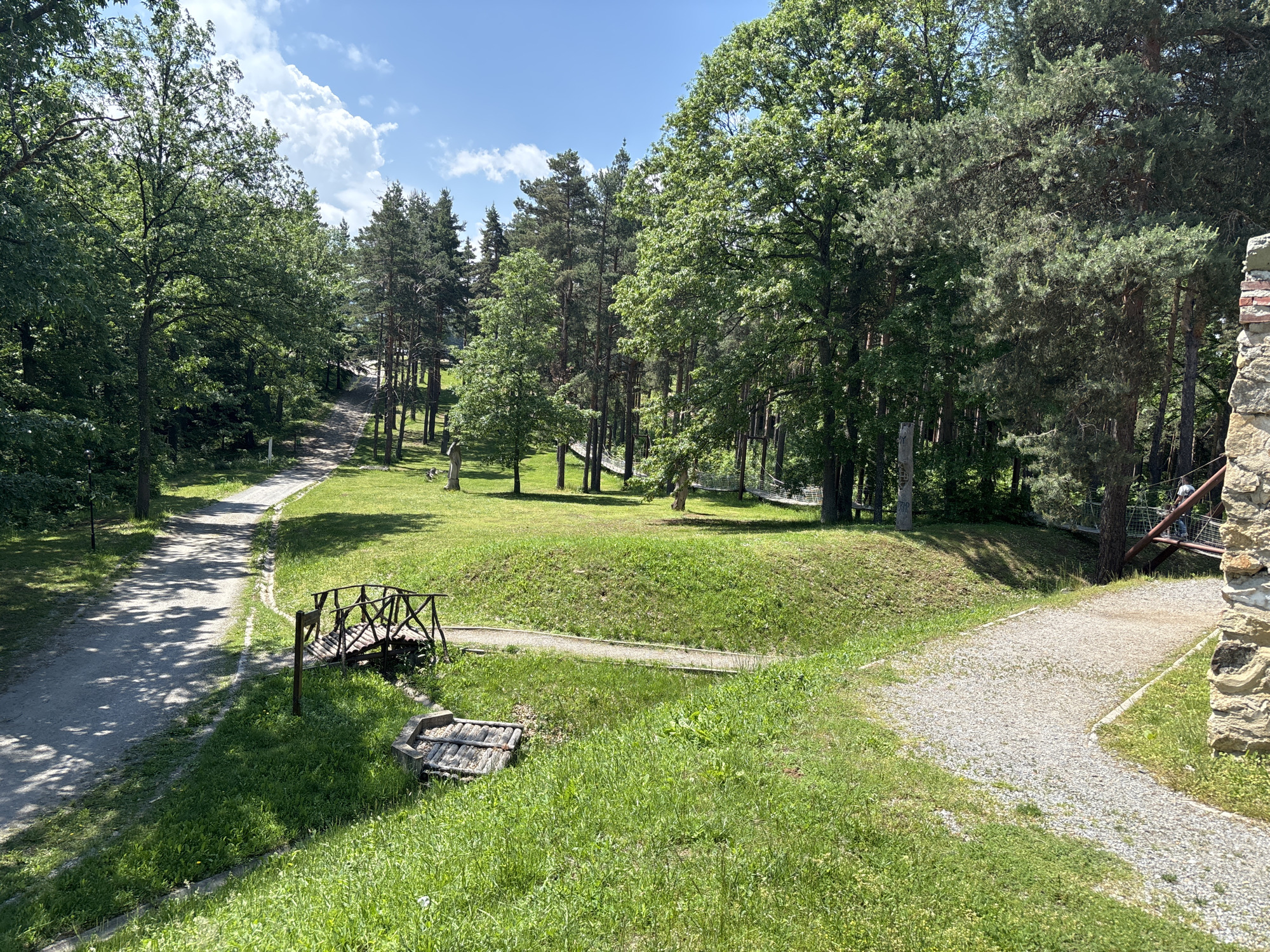 The image captures an outdoor recreational area within Belchinski Bani, Bulgaria, under a clear blue sky with scattered white cumulus clouds. The scene is dominated by lush green foliage from numerous deciduous and coniferous trees that form a dense forest in the background and along the edges of the frame. Sunlight casts distinct shadows across the landscape.
In the foreground and mid-ground, the ground is covered by vibrant green grass, interspersed with several unpaved paths. A winding dirt and gravel path originates from the left, extending into the mid-distance. A narrower, curvilinear gravel path runs horizontally in the lower-mid ground, leading to a small, dark-stained rustic wooden bridge with arched supports and cross-braced railings. Below and to the right of this bridge is a low, rectangular wooden structure, possibly a well cover, constructed from horizontal logs. On the far right, a partial stone wall or ruin, composed of irregular grey and brown stones with visible red brickwork higher up, occupies the foreground.
Beyond the grassy areas and paths, elements indicative of an adventure park are visible. A metal platform or support structure is positioned within the trees on the far right, and one individual wearing dark clothing is standing on this structure, facing away from the viewer. Faintly discernible in the mid-background are rope or net bridges strung between trees, suggesting a ropes course. The overall environment conveys a natural setting adapted for recreation.