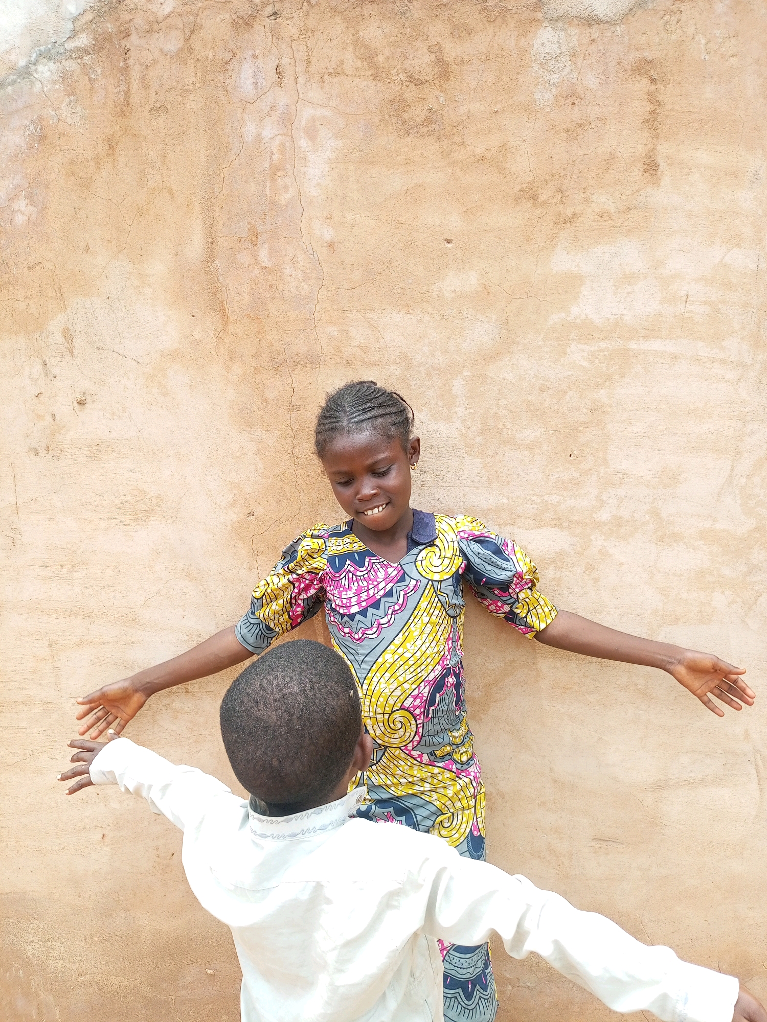 Two children are standing in front of a tan, textured wall in Tempure, Nigeria. The child on the right is a girl with braided hair, wearing a colorful dress with a blue collar and puffy sleeves. She is smiling and looking down. The child on the left is a boy wearing a white shirt, facing away from the camera. Both children have their arms outstretched. The wall behind them is weathered and cracked, suggesting an outdoor location.