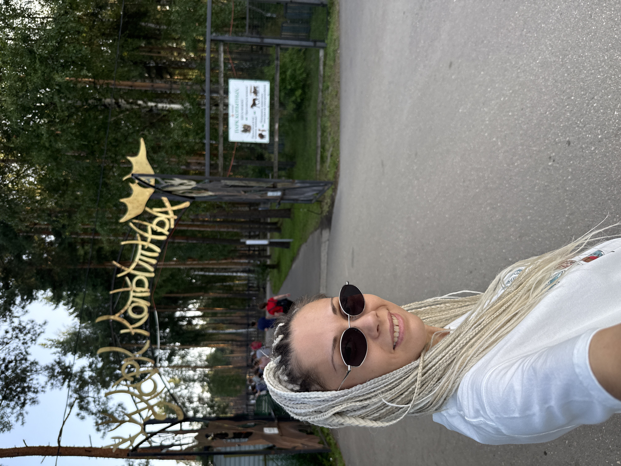 This is a vertical, eye-level, medium shot of a young woman taking a selfie. She has blonde braids that extend down her back and is wearing sunglasses and a white t-shirt. She is smiling at the camera, appearing happy. The background shows a park-like setting with tall trees and a paved pathway. A metal gate with a sign is visible, and ornate golden lettering adorns the entrance to the park. The sign appears to be in Cyrillic script and reads "ПАРК КОПЫТНЫХ", which translates to "Hoofed Animals Park". The time of day seems to be late afternoon or early evening, as indicated by the lighting and long shadows. The weather appears to be pleasant, with the sun casting a warm glow. The overall impression is that of a cheerful visit to a nature park or zoo.