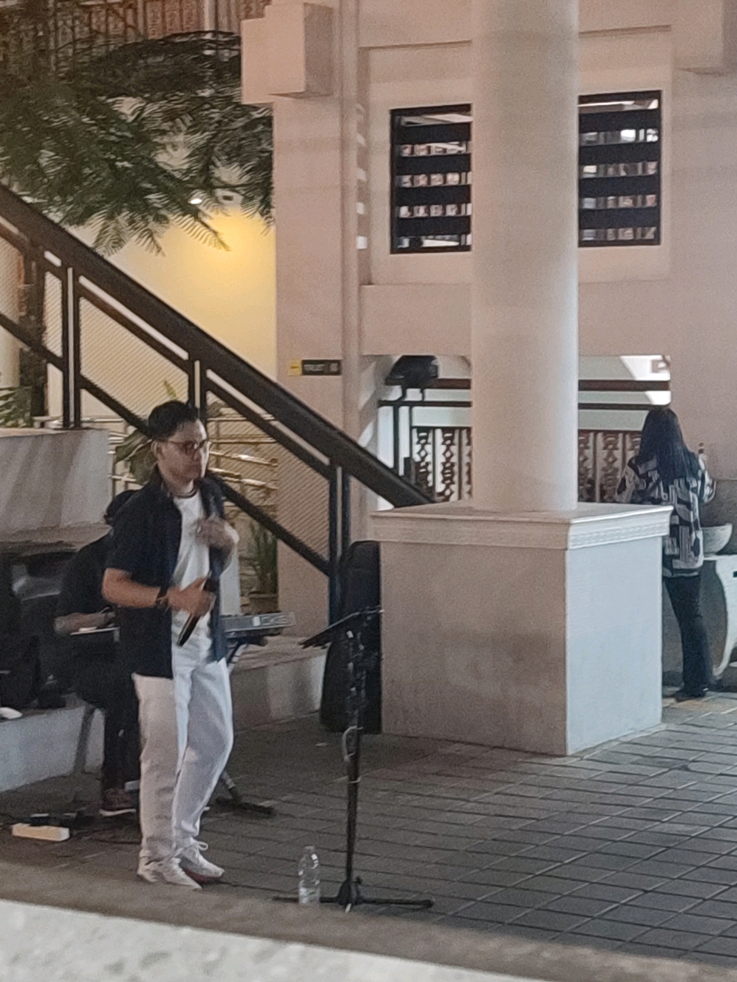 The image depicts an outdoor setting at night, likely in a public space in Yogyakarta, Indonesia. A man in his late twenties, wearing glasses, a dark unbuttoned shirt over a white t-shirt, and light-colored pants, is holding a microphone and appears to be performing or speaking. He is standing on a tiled surface, with a keyboard and a music stand visible behind him. To the right, a woman with long dark hair is standing, facing away from the camera, and appears to be holding a small object. In the background, there is a multi-level structure with stairs, balconies, and architectural features typical of urban Indonesian design, including decorative windows and columns. A large white pillar is a prominent feature in the mid-ground. The overall ambiance suggests a casual evening event or performance.