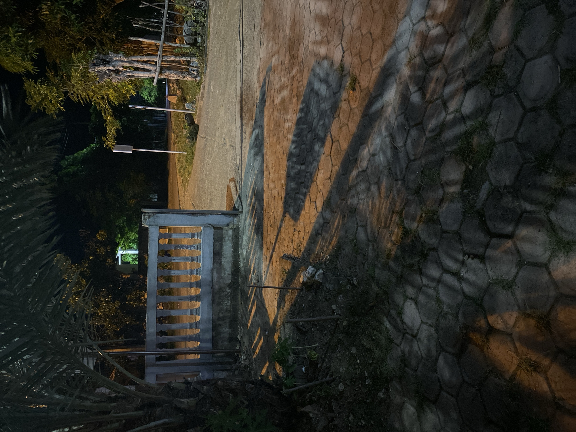 The image depicts an outdoor scene at night, likely in the city of Lambaro Kueh, Indonesia. The foreground is dominated by a paved area with hexagonal interlocking concrete pavers, interspersed with patches of grass and dirt. A stone fence or low wall is visible along the edge of the paved area. To the left, a portion of a palm tree trunk is present, and further back, an open gate with a decorative balustrade made of concrete pillars and horizontal bars stands. Beyond the gate, a dirt or gravel pathway leads into a darker area with vegetation. In the background, trees are visible, some illuminated by artificial light, creating distinct shadows on the ground. A white sign on a pole is situated in the mid-ground to the right. The overall lighting suggests artificial illumination from an unseen source, casting long shadows and highlighting textures.