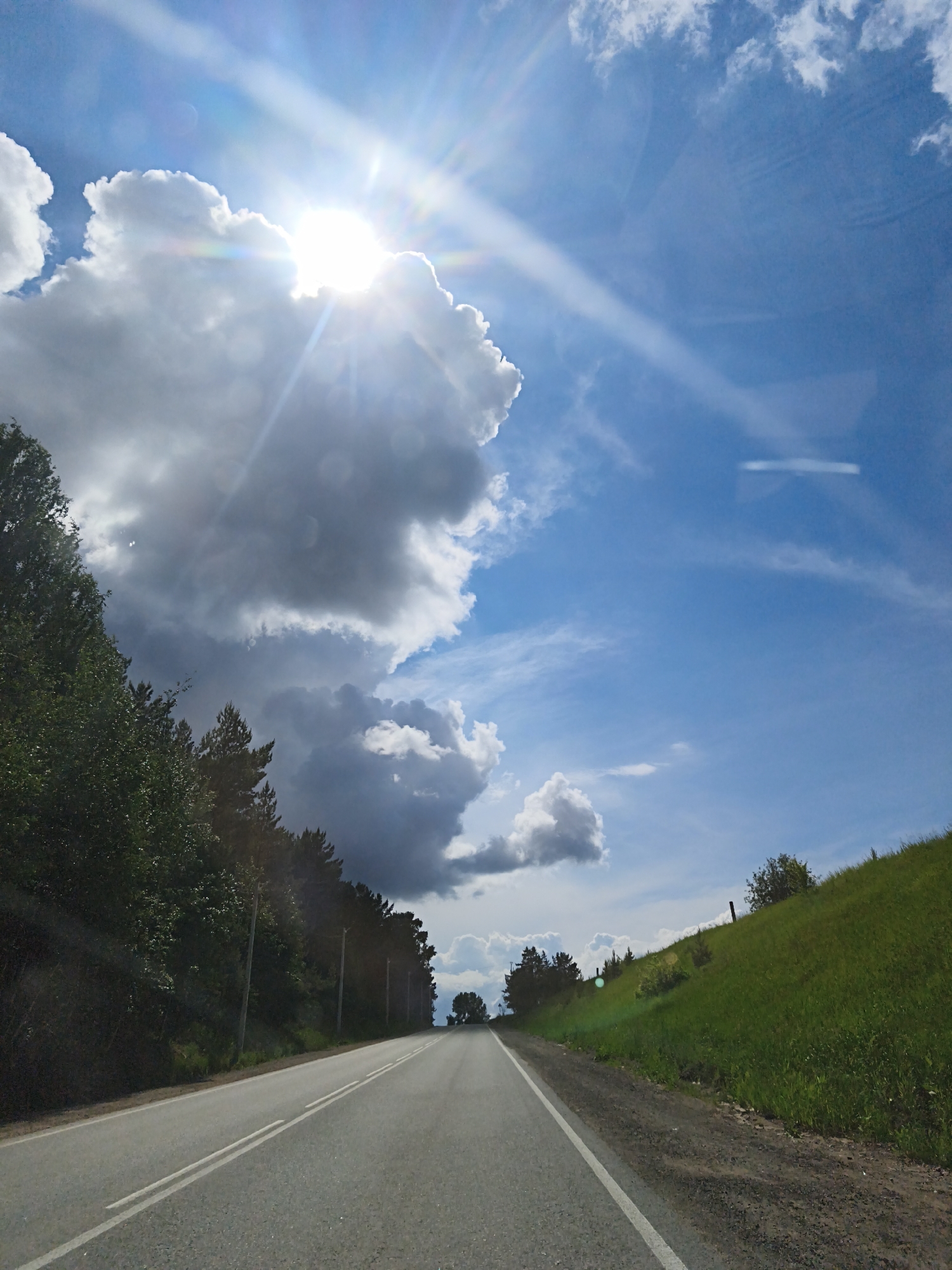The image depicts a road leading into the distance. On the left side of the road is a line of trees, and on the right is a grassy hill. The sky above is blue with bright clouds, with the sun obscured by the clouds but still emitting rays of light. The location is potentially near Chaikovsky, Russia, considering the vegetation and road conditions observed, but further confirmation is required for verification. No people or animals are visible in the image. The road has two white lines delineating the lanes. There are no other vehicles visible on the road. The environment is rural or semi-rural.