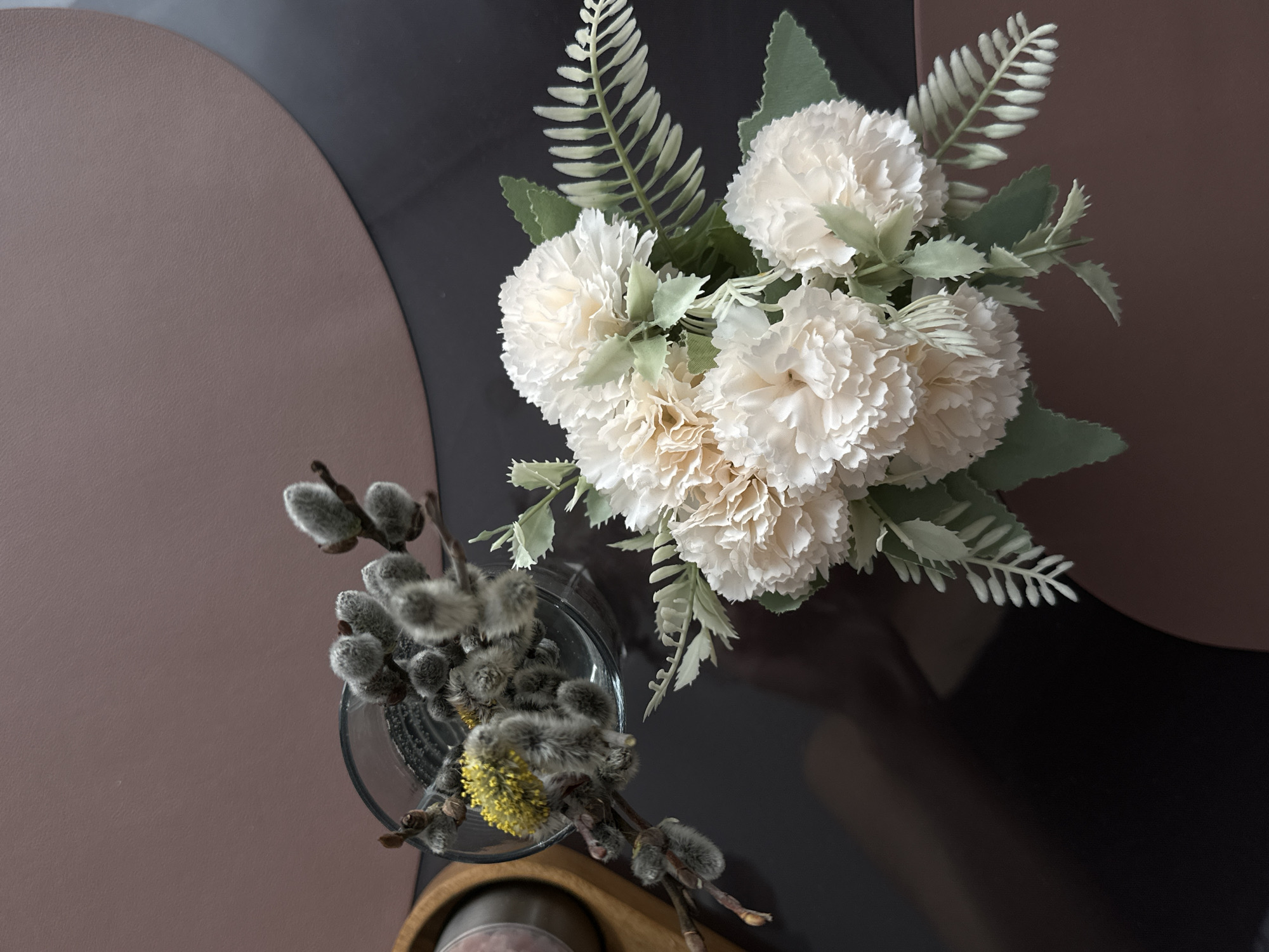 A high-angle shot depicts a table setting with a floral arrangement and willow branches. The table's surface is a two-tone design, featuring a matte brown semicircle and a glossy black semicircle. A bouquet of white pompom-like flowers with green and silver foliage is positioned in the upper right quadrant of the frame. To the lower left, a glass vase holds several willow branches with fuzzy catkins, one of which displays a bright yellow bloom. The environment is clean and well-lit. There is no information to indicate the location of the image is in the city of Подшивалово, Russia.
