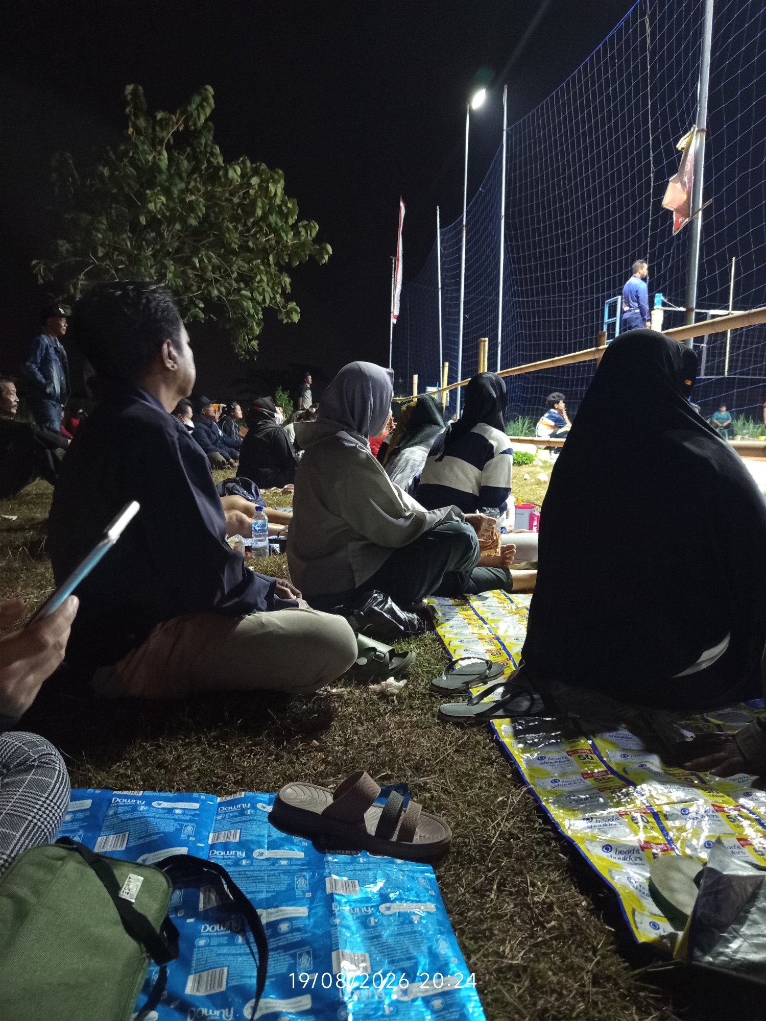 The image shows a group of people sitting on the ground at night, likely watching an event. The scene appears to be in Canggu, Indonesia, as indicated by the timestamp in the lower right corner. The ground is covered with grass and what seem to be tarps or mats, some with branding visible, including "Downy" and "Head & Shoulders".
In the foreground, several individuals are seated. One person on the left is wearing a dark jacket and light-colored pants, and has a stylus or pen in their hand. Another person in the center is wearing a grey hooded jacket, and next to them, a person is wearing a black hijab and a black outer garment. Further back, other people are visible sitting on the ground, some wearing headscarves.
Behind the seated crowd, a volleyball net is set up, illuminated by lights, suggesting a game or competition is taking place. Two figures are visible on a platform behind the net, one in a blue shirt. A tree with green foliage is on the left side of the image, and several flags are attached to poles near the net.
The overall impression is of an informal outdoor gathering, possibly a local sporting event or cultural activity in a community setting in Canggu.