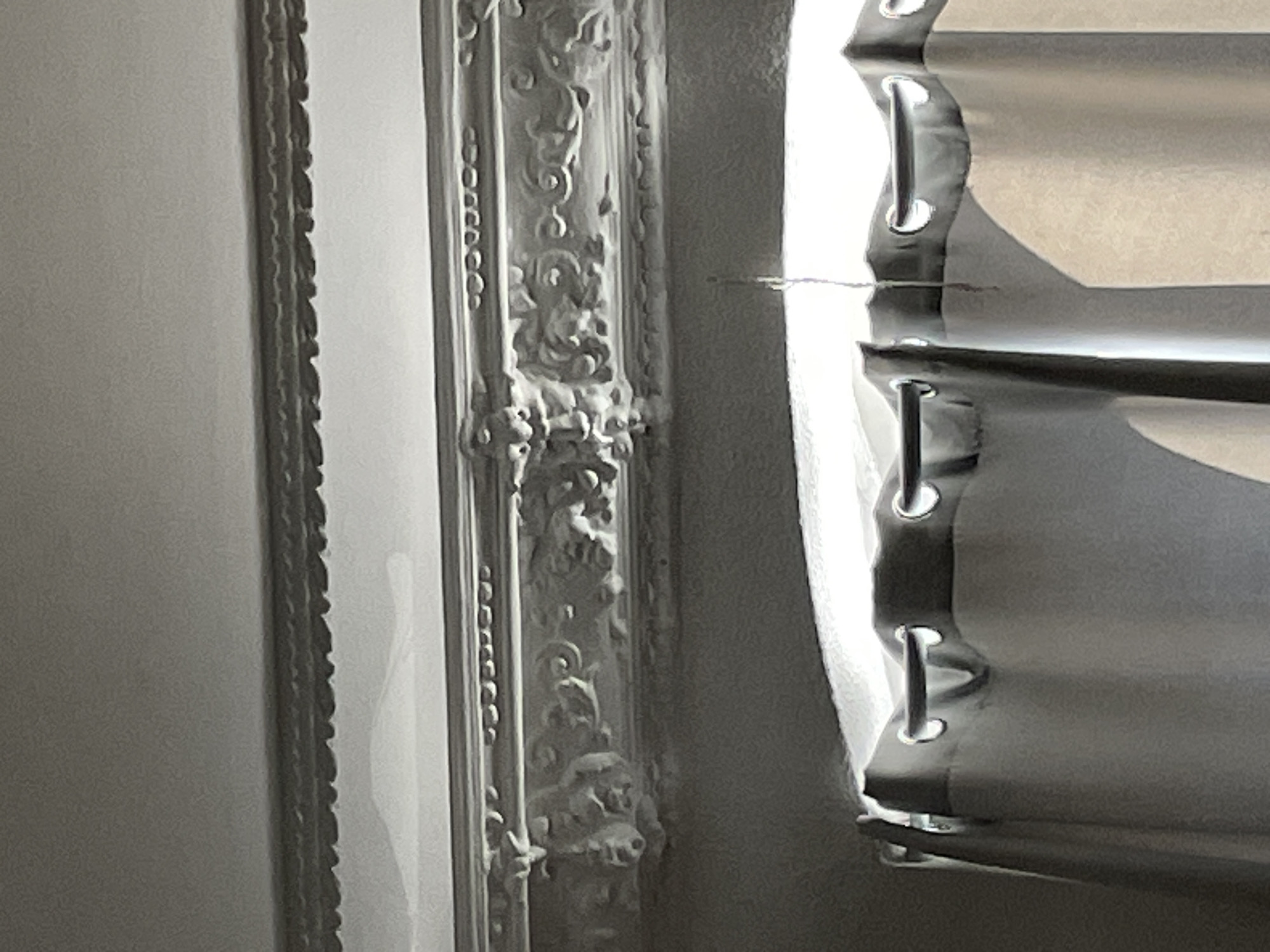 The image shows a close-up view of an ornate ceiling molding with carved details. Below the molding, gray curtains are partially drawn, obscuring a window. The curtains are hung from a dark rod with grommet holes. A thin, dark string hangs down from the ceiling area in the center of the image, in front of the curtains. The lighting suggests an indoor setting. The scene is devoid of people or any discernible outdoor elements. The location is identified as Unknown, Unknown.