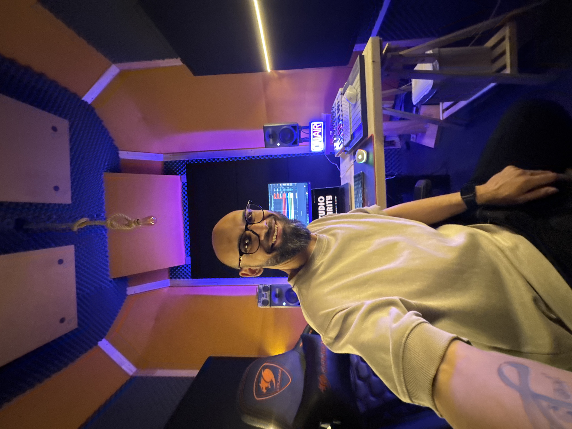 A man with a shaved head and beard sits in a recording studio. He wears glasses and a light-colored sweater, and a watch on his left wrist. He smiles at the camera while seated in a black and orange Cougar gaming chair.
The recording studio features acoustic foam panels in blue, orange, and black. A computer screen displaying audio waveforms and the words "Studio Priority" is visible. Speakers and an "ON AIR" sign are mounted on the wall. A mixing board and other audio equipment sit on a shelf above the computer monitor. A rope hangs from the ceiling.
The location is identified as a recording studio in Plovdiv, Bulgaria.