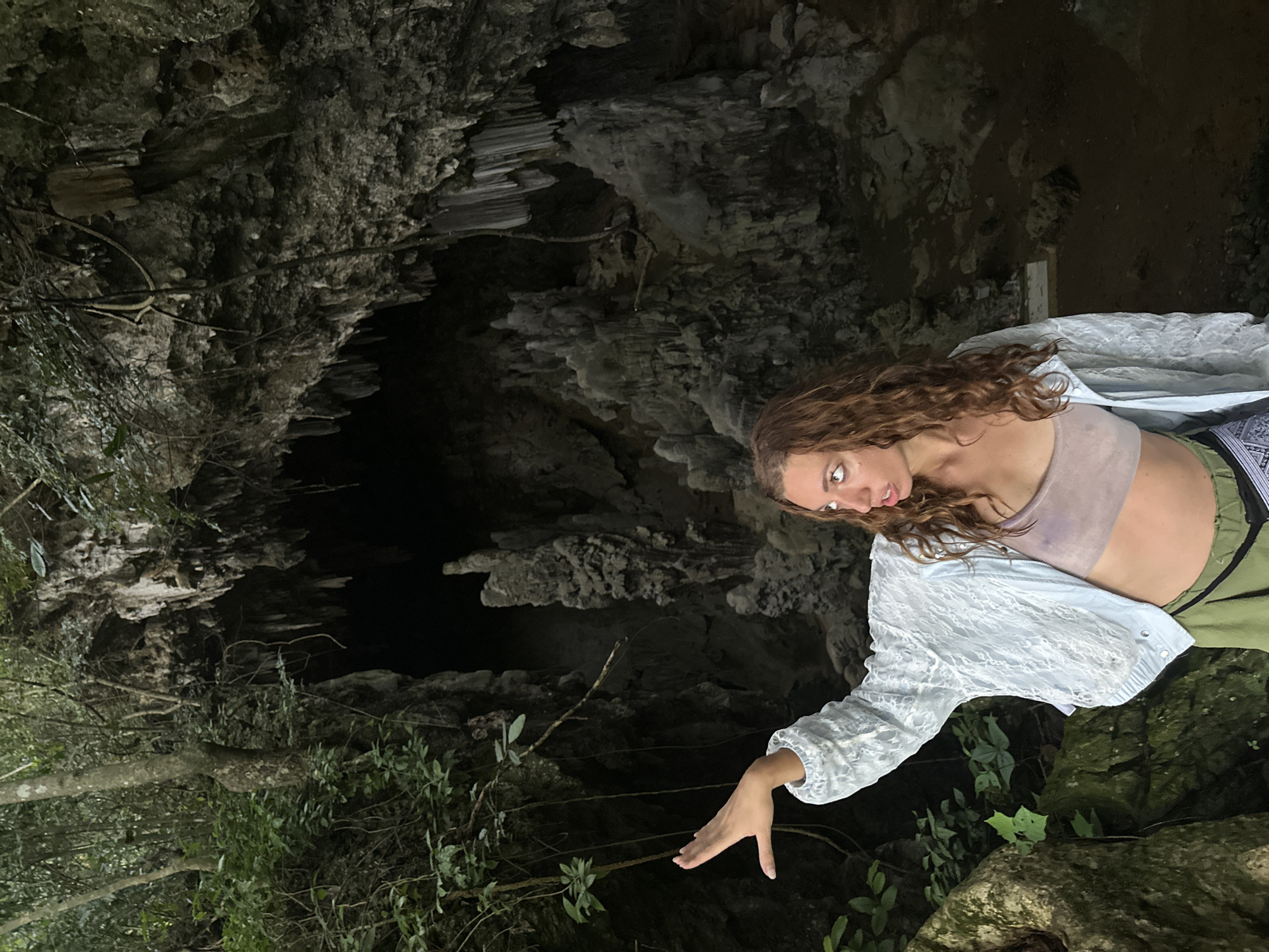 A woman with long, curly brown hair stands in front of the entrance of a cave. She is wearing a light blue lacy jacket, a beige crop top, and green pants. Her left arm is extended upwards and to the left, with her hand open. She is looking to her left, with her mouth slightly open as if speaking or surprised. The cave entrance is large and dark, with stalactites hanging from the ceiling. The surrounding environment is lush and green, with trees and foliage visible. The ground appears to be rocky and uneven. The overall scene suggests a natural, outdoor setting, possibly a tourist attraction within a rural area.