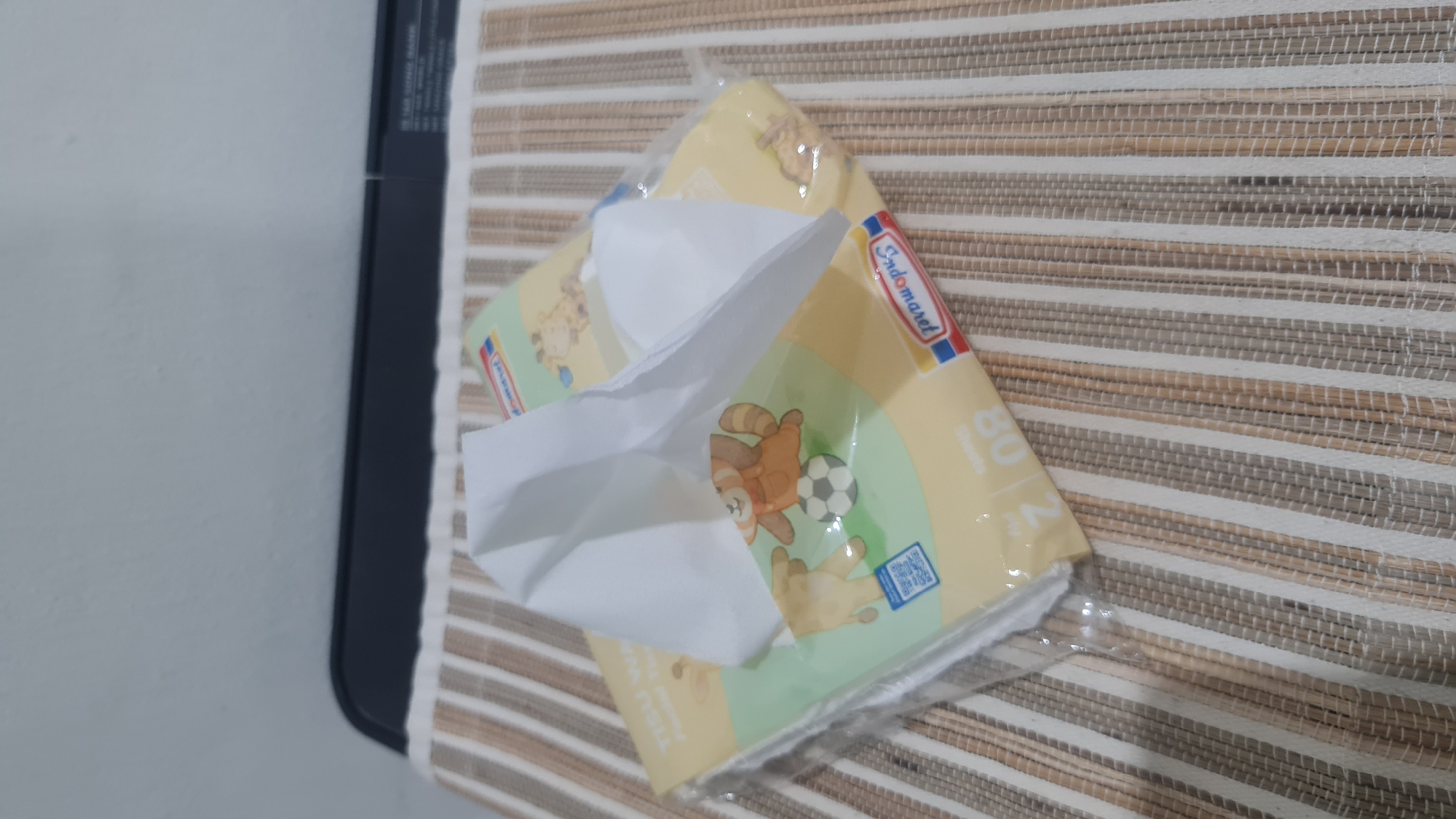A pack of Indomaret brand facial tissues is placed on a striped placemat. The tissue pack has a yellow background with cartoon animal illustrations, including a red panda playing soccer and a giraffe. The words "TISU WA" and "Indomaret" are visible on the packaging. A portion of a black electronic device, possibly a printer, is visible in the background above the placemat. The scene is indoors, and the lighting suggests it is daytime.