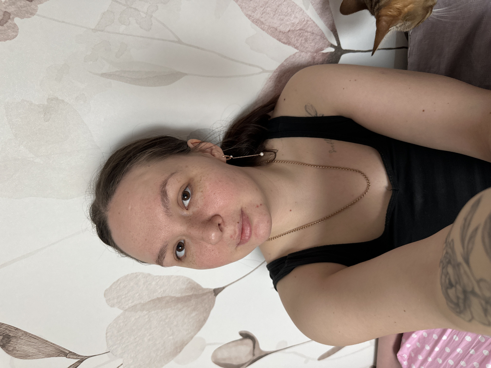 The image portrays a female individual and a domestic cat within an indoor residential environment in the city of Chaikovsky, Russia. The female, appearing to be in her 20s or 30s, occupies the center of the frame, facing forward with a neutral expression, looking directly at the camera. She possesses light brown hair, dark eyes, and a complexion with visible freckles and some erythema on the cheeks and nasal area. She is dressed in a black tank top and accessorized with a gold-colored chain necklace and a gold-colored dangle earring featuring a pearl in her right ear. A small script tattoo is visible on her upper right chest/shoulder, and a larger tattoo design is partially visible on her left forearm. To the right, the head and upper torso of an orange-brown tabby cat are present, oriented downwards towards the female's lower left arm area. The background consists of a wall adorned with light-toned floral wallpaper depicting large, muted flower patterns. Below the female's left arm, pink fabric with white polka dots is visible, likely bedding. To the right, beneath the cat, a plain light purple/grey fabric is present, also consistent with bedding or soft furnishings. The scene is illuminated by even, ambient indoor lighting.