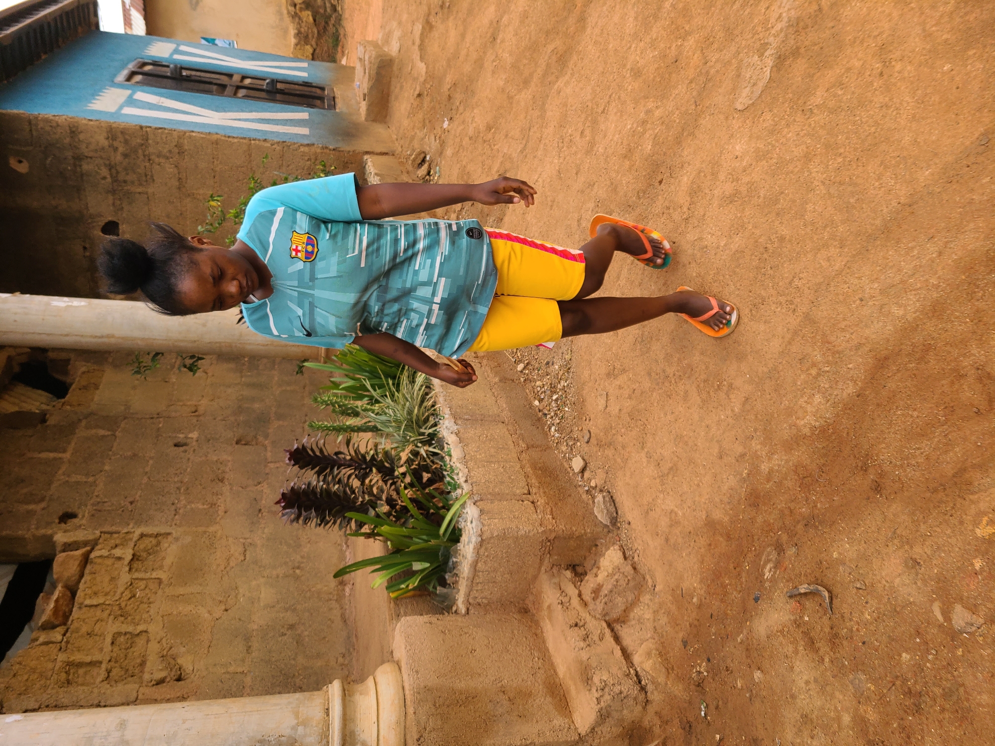 A young person wearing a turquoise and white FC Barcelona shirt, yellow shorts, and orange flip-flops stands on a reddish-brown dirt surface. A weathered building with a blue section and white trim is behind them. Plants are visible in front of the building's base. The location appears to be an outdoor area in a residential neighborhood in Jos, Nigeria, judging by the architecture and environment.