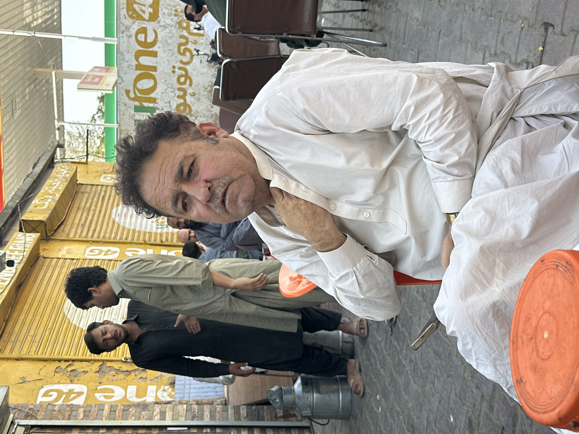 An outdoor medium shot captures a man seated in a red chair in Quetta City Tehsil, Pakistan. The man, of middle age with dark, curly hair and a mustache, wears a light-colored shalwar kameez. His right hand rests on his chest, and his left hand is clasped over his lap. To his left, in the background, two men in traditional clothing stand near a yellow building with a shuttered entrance and signage. Further back, other individuals are visible, some seated, near tables and chairs. The environment appears to be a street or marketplace, with a metal barrel on the left and a bright orange circular object in the foreground on the man's lap. The overall scene suggests a casual gathering or waiting area.