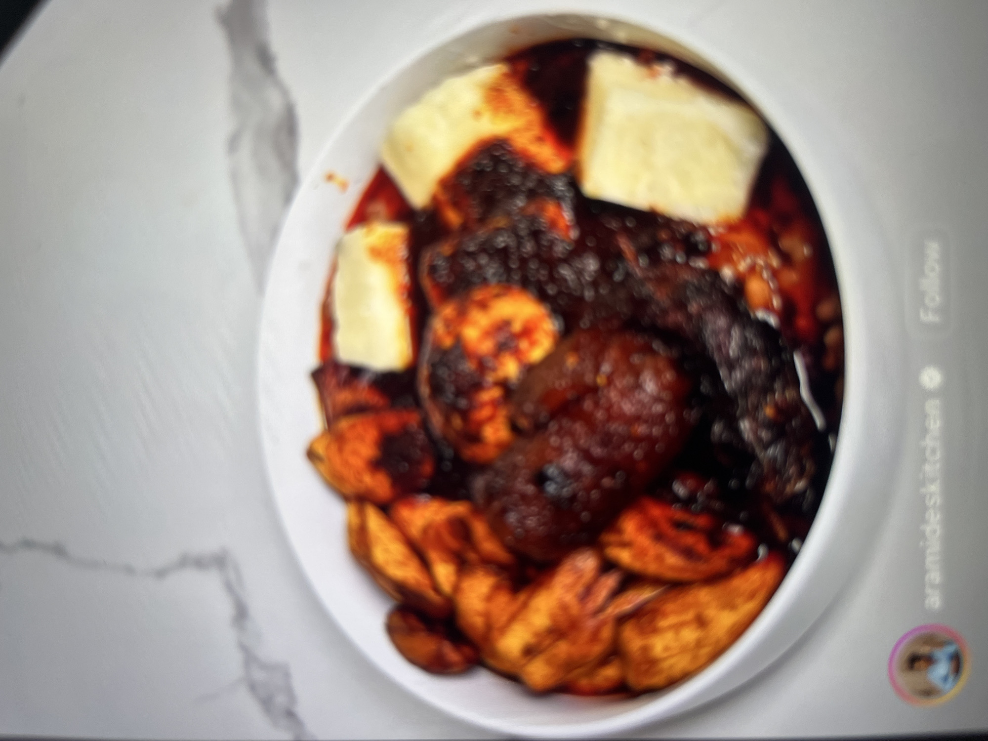 A close-up, blurry overhead shot shows a white bowl filled with a dark, reddish-brown stew. The stew contains visible pieces of fried plantains, and several light-yellow, square-shaped pieces, possibly yam or cheese, are submerged in the sauce. The bowl sits on a light-colored, marble-patterned countertop. In the bottom left corner, a profile picture of a person is visible, along with the text "aramideskitchen" and a "Follow" button, suggesting this is a social media post about food. The image does not provide any information to determine its location in Abuja, Nigeria.