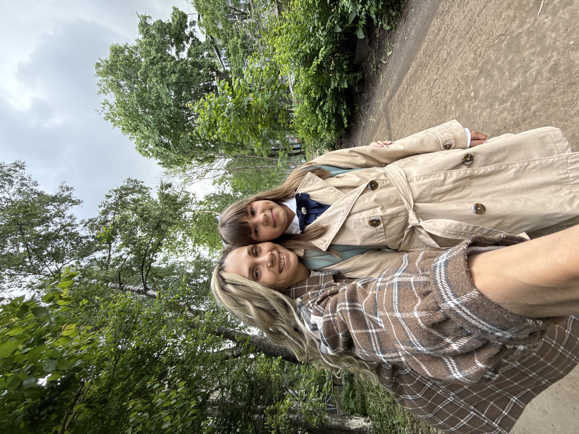 A woman and a girl stand together on an asphalt path lined with green vegetation. The woman wears a brown and white plaid shirt and has long blonde hair. The girl wears a beige trench coat over a white collared shirt with a dark blue bow. The background shows a cityscape visible through the trees, beneath an overcast sky. The location is Шебунино, Russia.