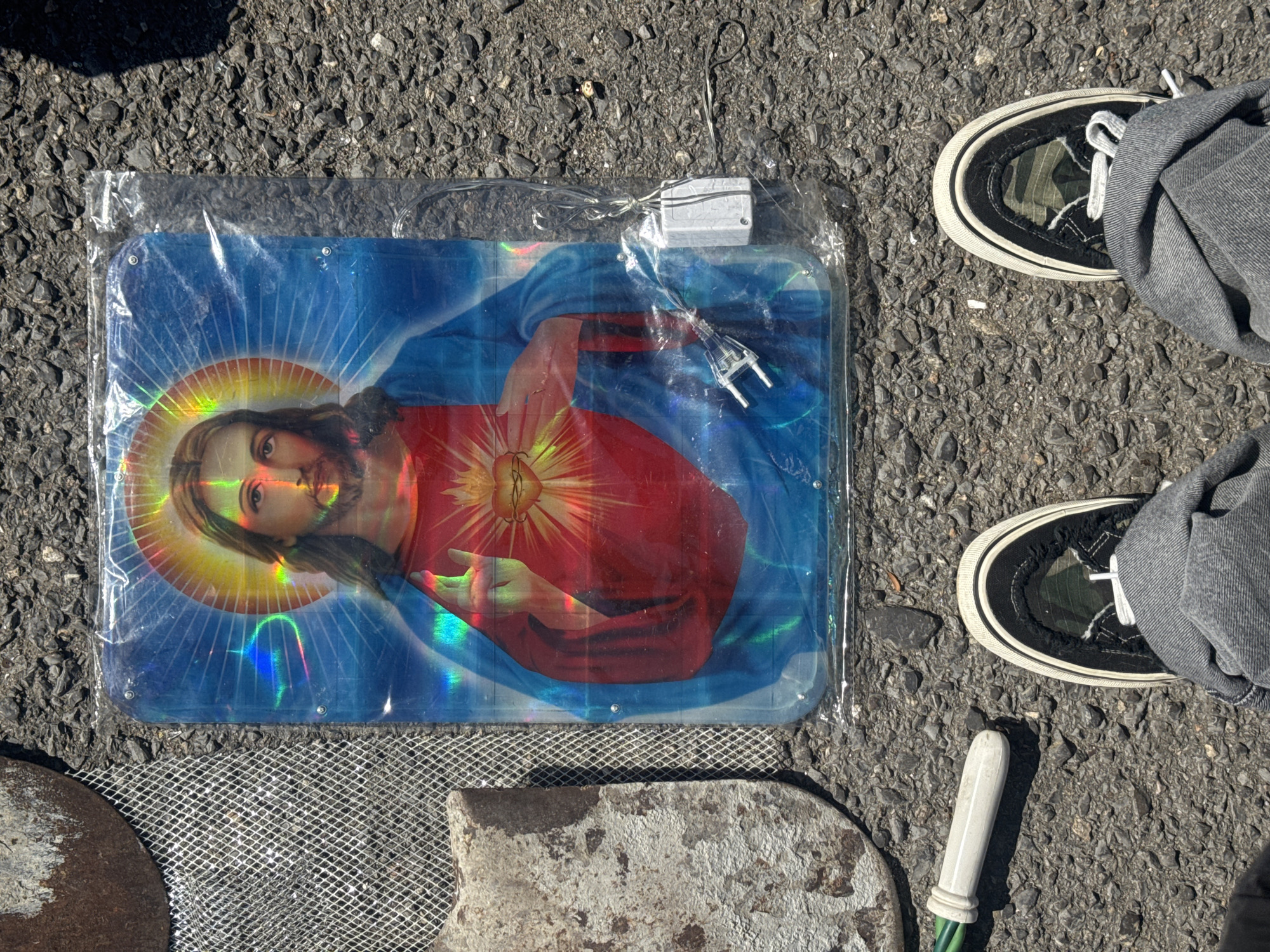 The image shows a person's feet wearing grey jeans and black and white sneakers, standing on an asphalt surface. In front of the person is a rectangular plastic-covered image of Jesus with a glowing heart, illuminated by a string of small lights. To the left of the image are some metallic mesh and concrete objects. The scene appears to be outdoors, possibly in a yard or street, given the asphalt and the sunlight casting shadows. There is no indication of the specific location within the city of Sofia, Bulgaria.