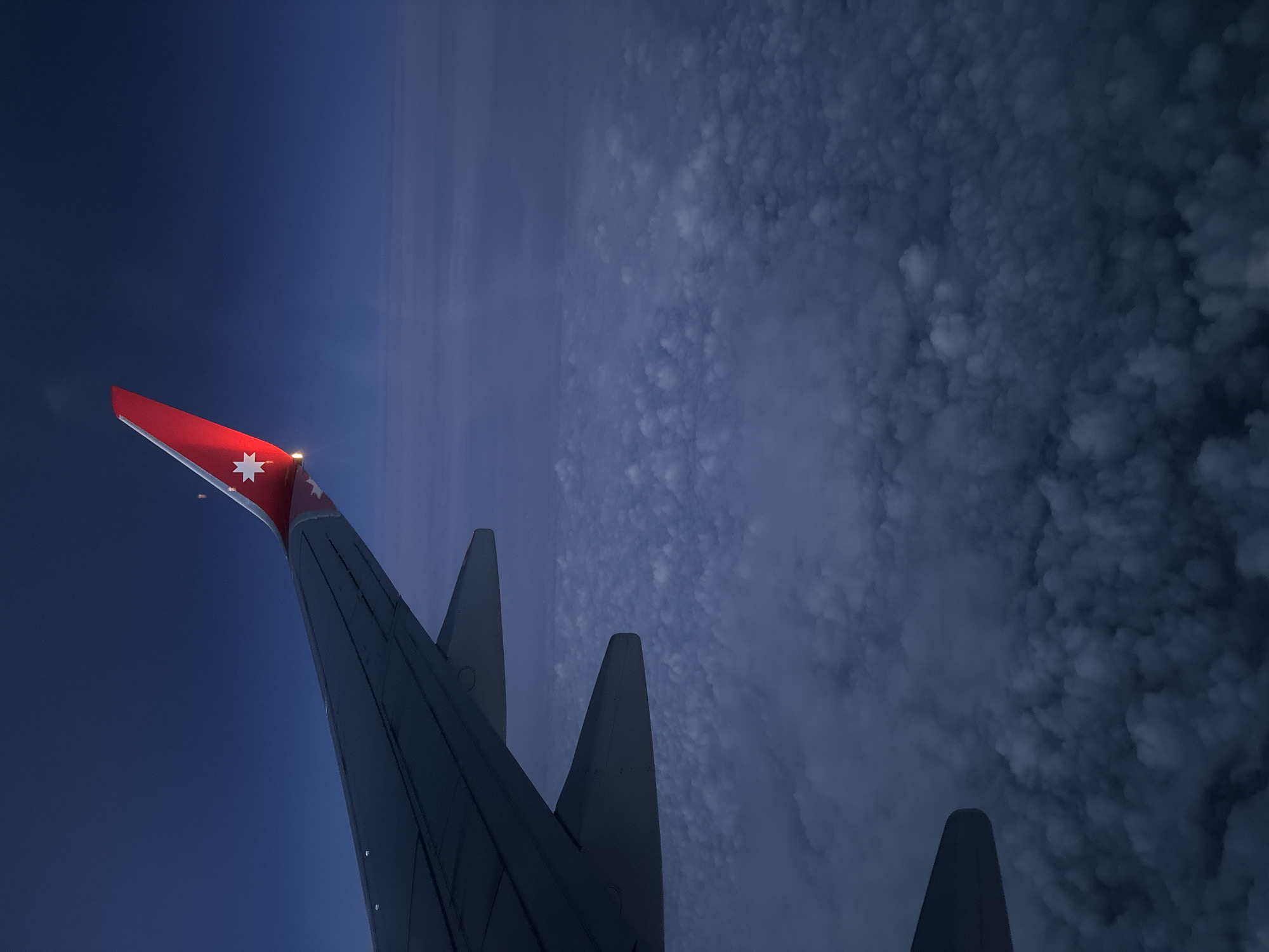 A view from an airplane window shows the plane's wing and a landscape of clouds or terrain. The wing has a red tip with a white star emblem, indicating it might belong to an airline from Australia or a similar region. The sky to the left of the wing appears dark, possibly suggesting nighttime or low light conditions. The right side of the image shows a dense, textured surface, likely clouds seen from above. The composition is tilted, giving a dynamic perspective. The location is not specifically identifiable as Морозовка, Russia, from the visual information provided.