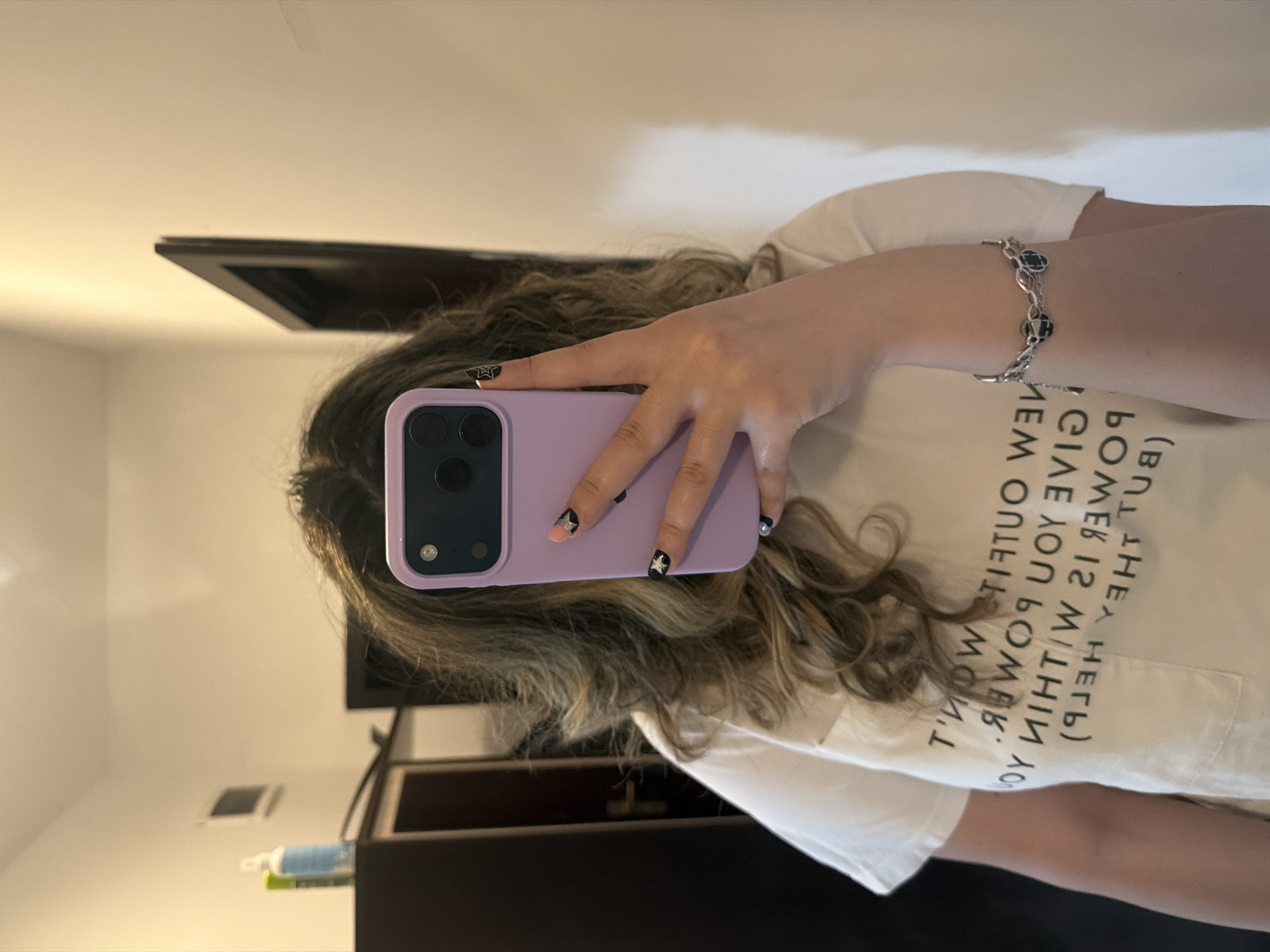 This image is a mirror selfie taken indoors in Plovdiv, Bulgaria. It features a person's right arm, hand, and the upper part of their head with long, wavy brown hair. The person is holding a light purple smartphone, encased, with its triple-camera setup visible, implying it's an iPhone Pro model.
On the person's right wrist, a silver chain bracelet with black, possibly clover-shaped, charms is visible. Their fingernails are painted with a distinct style: some are black with gold or silver star decals, while others have a natural or light pink base. The person is wearing a light-colored t-shirt with mirrored text, which clearly reads, "BUT THEY HELP GIVE YOU POWER IS WITHIN." The surrounding text is partially visible but less legible.
The background, seen through the mirror, consists of light-colored walls. Reflected in the upper part of the mirror is a dark-framed rectangular object, possibly a picture frame or a television screen. In the lower left, some toiletries (light blue and green bottles) are visible on a darker surface, likely a shelf or cabinet. A square vent is also visible on the wall. The lighting is bright and even, suggesting an interior setting, possibly daytime or well-lit. The image is oriented horizontally due to being rotated 90 degrees clockwise from its original capture.
