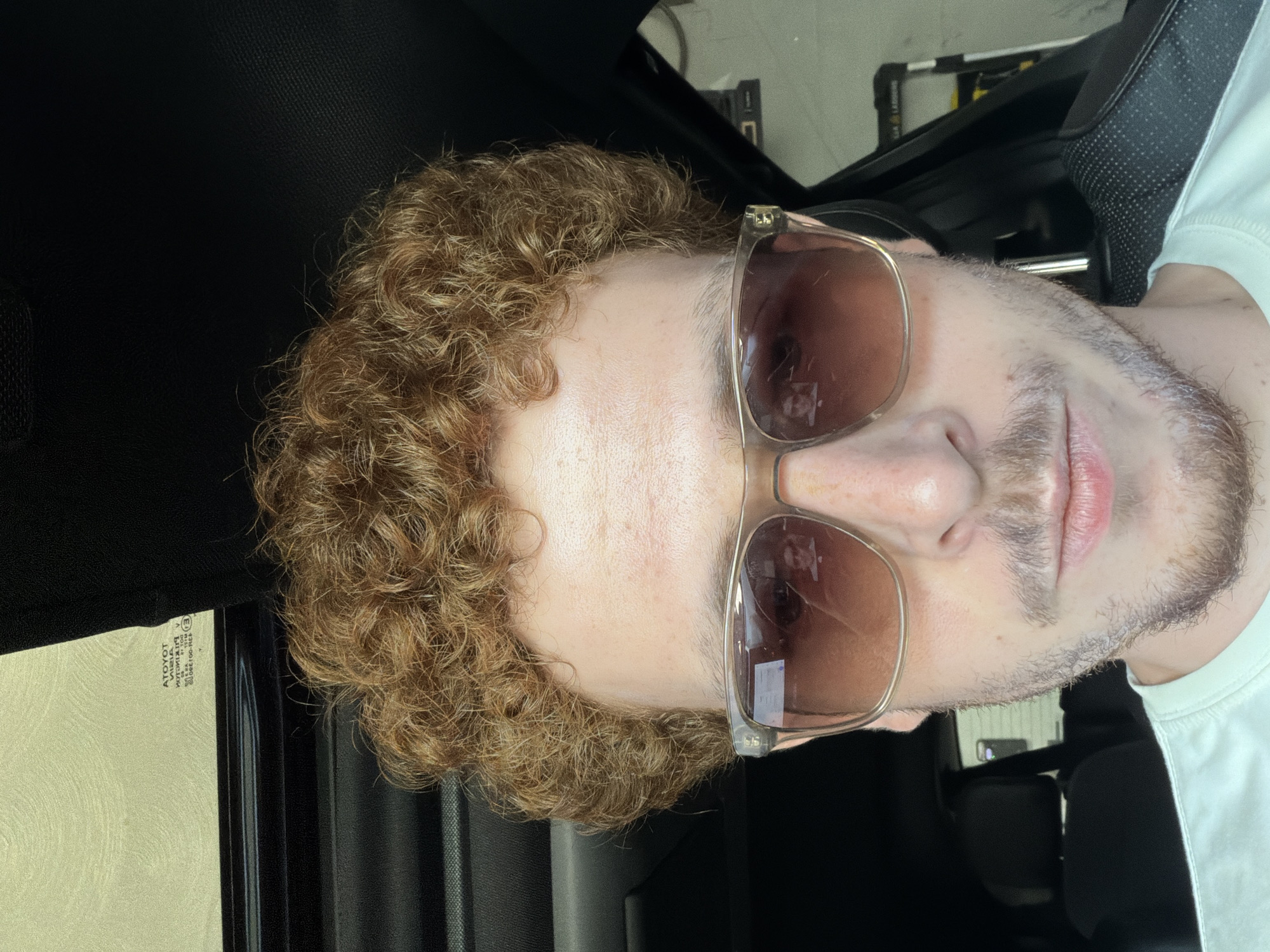A male individual with reddish-brown, curly hair, fair skin, and a light-colored mustache and goatee is depicted in a close-up, chest-up shot inside a vehicle. He is wearing sunglasses with light brown frames and tinted lenses, which reflect faint, distorted images of a person and interior details. His lips are closed. He wears a light-colored t-shirt. The immediate background shows the dark interior of the car, with visible black textured upholstery on the roof and pillars. Above, a sunroof is partially open, admitting light. To the left, a car window reflects an exterior building with light-colored siding and contains etched manufacturer information, including "ATOYOT" (Toyota reversed) and various regulatory symbols such as "V" and "E". Dark car seats are visible in the rear of the cabin, and elements of the car's door panel are present on the left side of the frame. The precise location within Pepper Pike, United States of America, is not identifiable from the visual content.